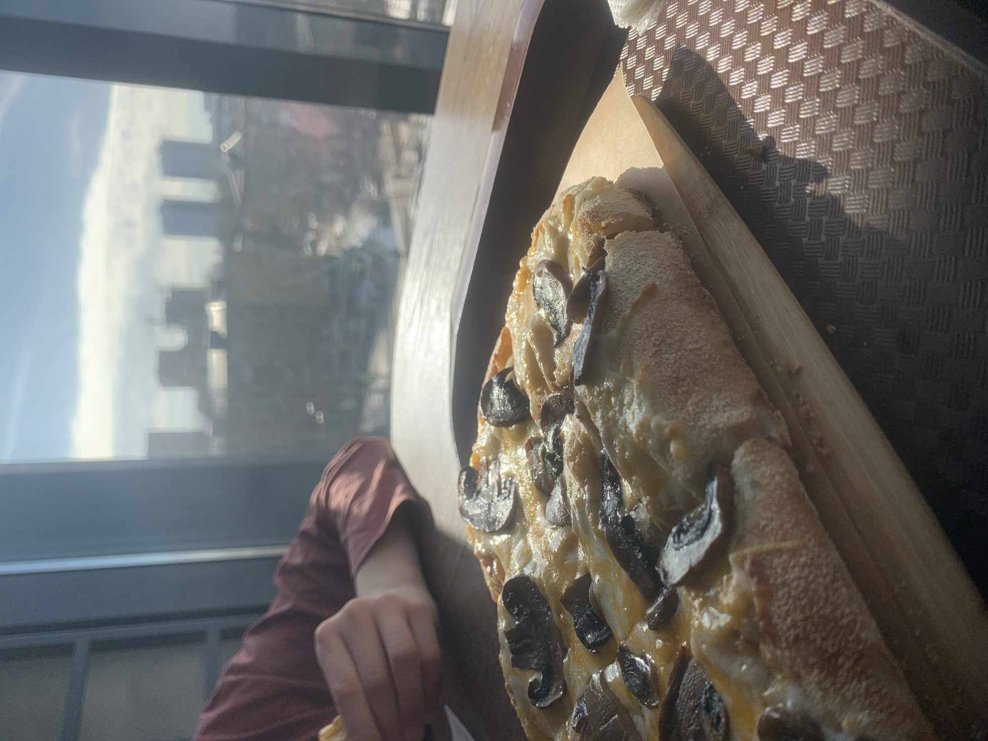 An indoor scene on an upper floor, likely a restaurant or cafe, overlooks a sunny cityscape in Izhevsk, Russia. In the foreground, a rectangular pizza, topped with melted cheese and sliced mushrooms, rests on a light wooden serving board positioned on a dark, patterned placemat. A stack of white napkins is visible to the far right on the placemat. To the mid-left, the forearm and hand of a person, clad in a reddish-brown long-sleeved shirt, are visible, holding a piece of food, indicating an action of eating. The table surface is dark wood. In the background, a large window reveals a bright blue sky with faint clouds. Through the window, a blurred panoramic view of Izhevsk includes several indistinct buildings. Two prominent, slender high-rise buildings, appearing as a pair, are visible in the central-right distance, characteristic of a modern urban development within the city. Sunlight streams through the window, casting reflections and highlights on the table and food.