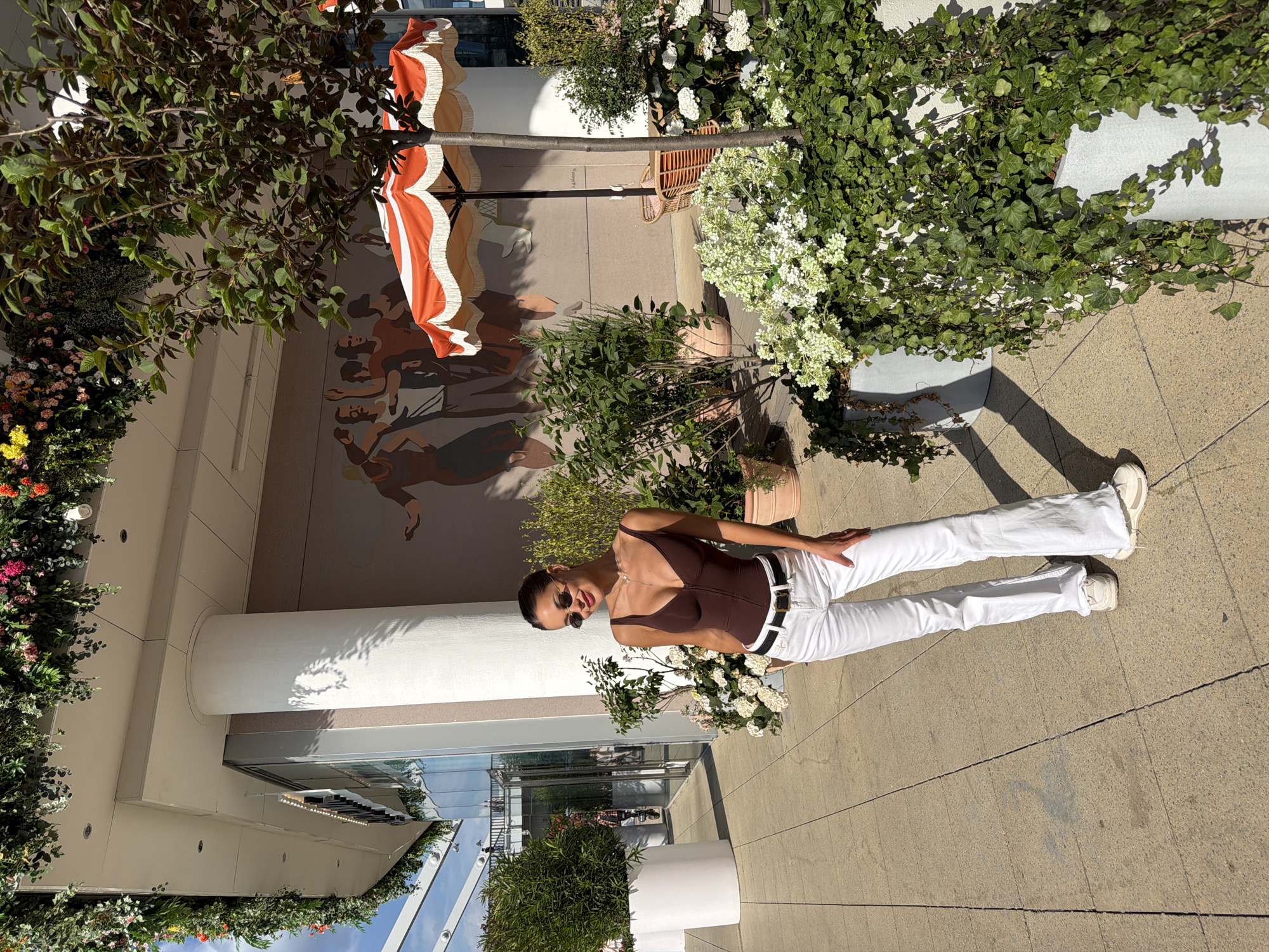A stylish young woman poses for a photograph outdoors in what appears to be a modern urban plaza or courtyard in Moscow, Russia. She stands towards the right of the frame, facing slightly left with a relaxed and confident posture. Her dark hair is pulled back, and she wears dark sunglasses, a form-fitting brown sleeveless top, high-waisted white pants, a black belt with a white buckle, and white sneakers. A delicate necklace is visible around her neck. She holds a bunch of white flowers in her left hand, which rests near her waist, while her right hand is casually placed on her hip.
The background features a light-colored building wall with a large mural depicting stylized figures in dynamic poses, suggesting movement or dance. To the left and slightly behind, a large orange and white striped umbrella with a fringed edge is partially visible. The scene is lush with greenery, including various potted plants with white and colorful flowers, and climbing ivy adorning planters. The ground is paved with light-colored tiles, and strong sunlight casts distinct shadows, indicating a bright, clear day, likely midday or early afternoon. The overall impression is chic and inviting, within a well-maintained public or commercial space. No discernible text is visible in the image.