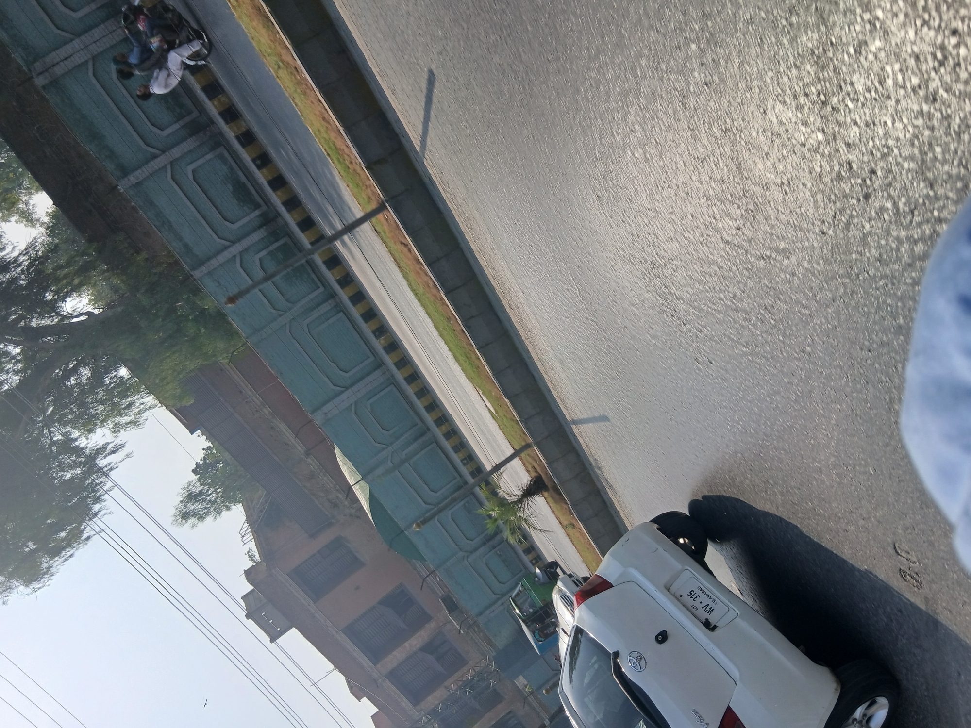 The image shows a street scene in Peshawar City Tehsil, Pakistan. A white Toyota Vitz is parked on the right side of the road. In the background, there is a light blue wall with a decorative pattern, and beyond that, a brick building. A green auto-rickshaw is driving on the road. Two people are riding a motorcycle in the distance. The road is paved and has yellow and black striped markings on the side. There are trees and power lines visible in the upper portion of the image. The scene is outdoors and appears to be during the daytime.