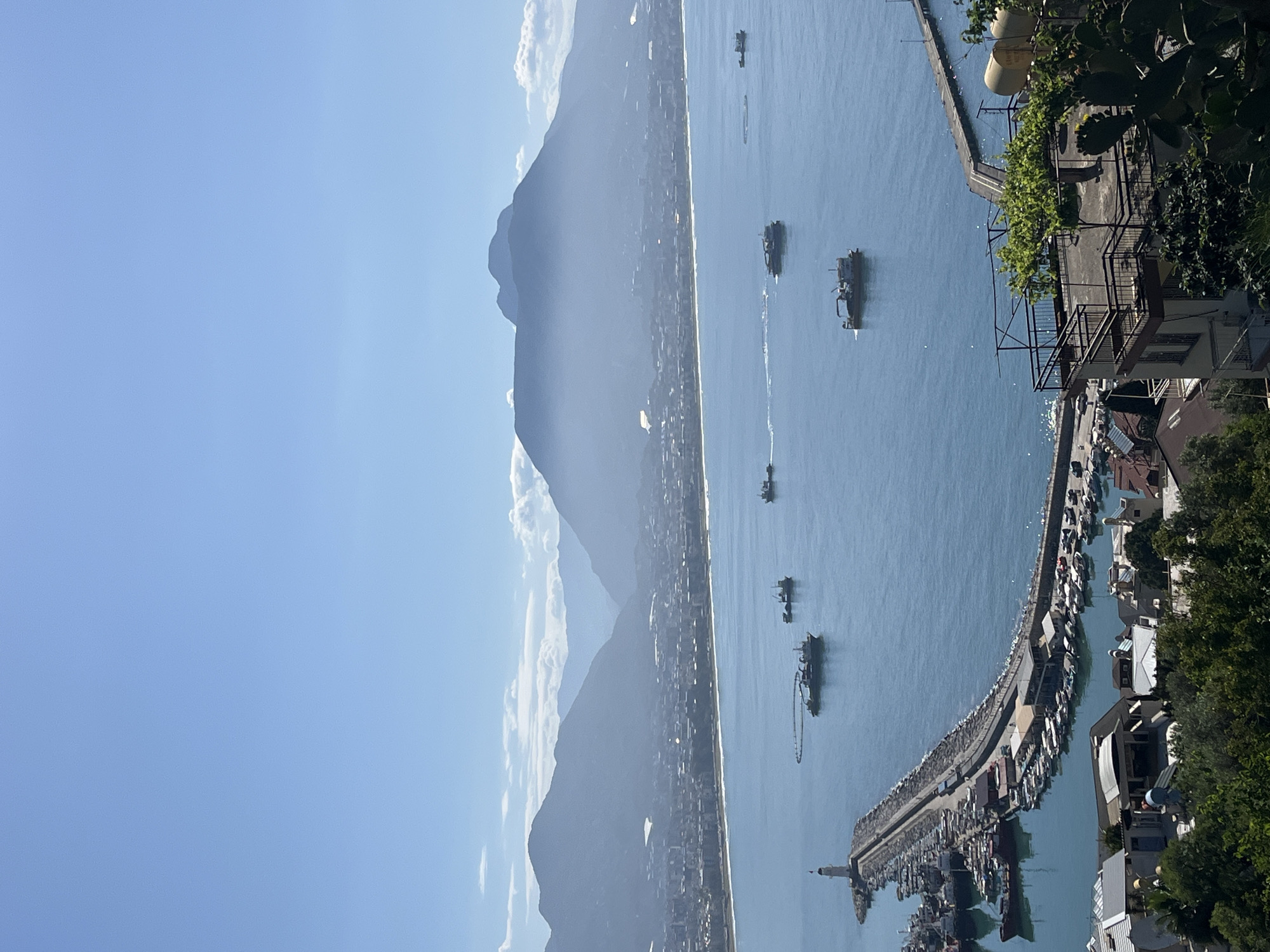 The image depicts a panoramic view of Alanya, Turkey, showcasing a harbor and surrounding landscape. The scene is set on a bright, sunny day with a clear blue sky dominating the upper portion of the frame. A mountain range rises along the left side of the image, partially obscured by a haze or light cloud cover.
A coastal city is visible at the base of the mountains, lining the shoreline. The sea occupies the central portion of the image, dotted with several boats of varying sizes. A harbor constructed from stone extends into the sea, hosting a multitude of smaller boats docked along its sides.
The bottom right corner of the image includes a view of buildings and greenery, suggesting a residential area. A building with a visible balcony and overgrown vegetation are present. A few other white roofs and building structures can be seen next to the vegetation.