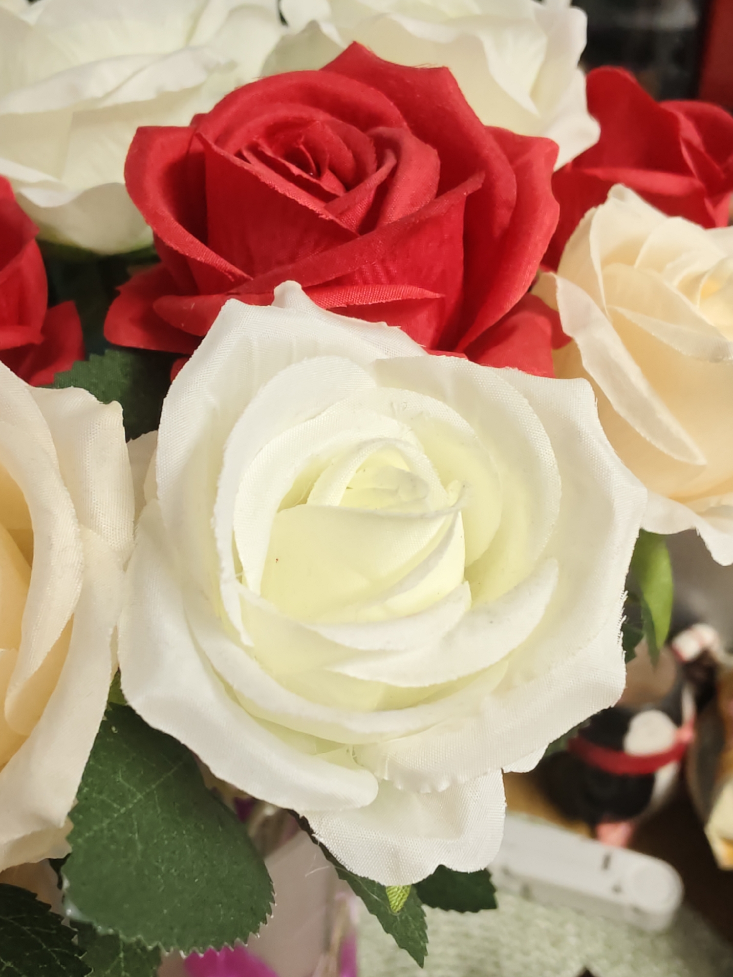 A close-up, overhead shot captures an arrangement of artificial roses in various colors, including red, white, and cream. The roses, with their fabric petals, are the primary focus, filling the frame. Some green artificial leaves are visible peeking through the blooms. In the background, out of focus, are indistinct objects, suggesting a decorative setting. The overall impression is of a static, artificial floral display. No information about Petah Tikva, Israel is present in the image.