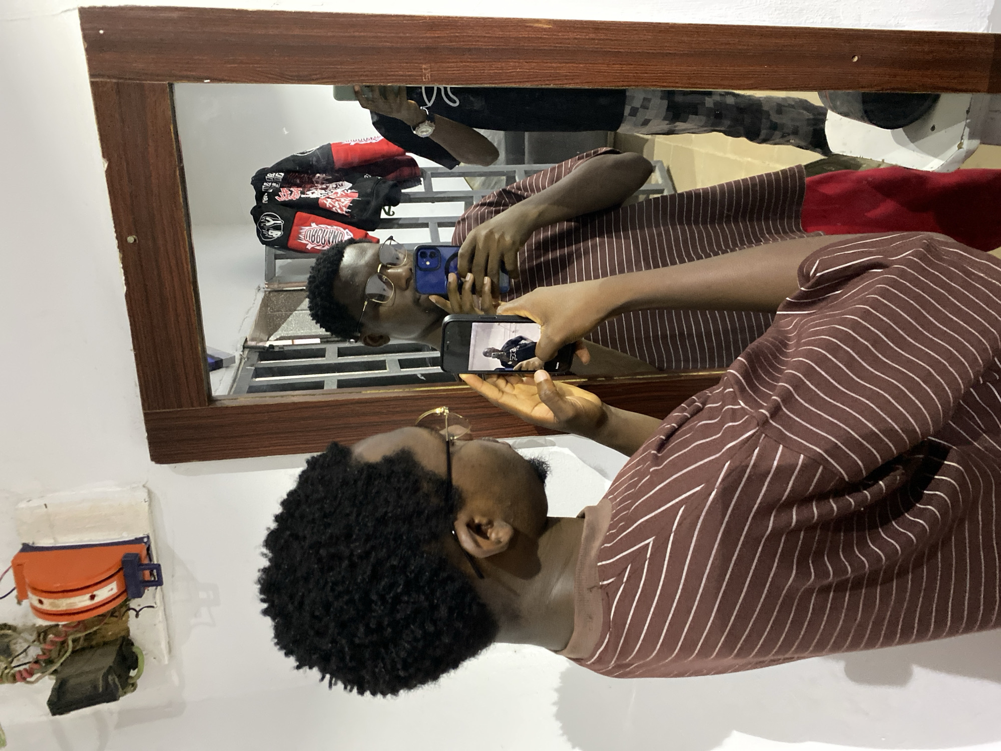 The image displays a young man taking a layered mirror selfie indoors. The original image appears rotated 90 degrees clockwise.
The subject is a man with dark skin, short curly black hair, wearing gold-rimmed glasses and a brown shirt with white vertical stripes. He is holding two smartphones. One phone, encased in blue, is held close to his face, partially obscuring his features. The second phone, with a black case, is held further out, aimed at a large mirror to capture his reflection. The screen of this black phone clearly shows an image of himself, creating an "image within an image" effect.
The mirror, set in a dark wooden frame, reflects the man and the room behind him. Visible in the reflection are a red and black patterned garment, possibly a jacket or hoodie, featuring some reversed, illegible text that resembles "OMABBD." Other items in the reflection include a dark patterned fabric (likely trousers) and a white object, suggesting a cluttered background like a personal wardrobe or storage space.
To the left of the mirror, on a white wall, an orange and blue electronic device with visible wiring is mounted, possibly an old intercom or part of an electrical setup. The scene depicts an informal, personal indoor environment, likely a bedroom or dressing area, under artificial lighting. No specific emotions are clearly discernible from the partially obscured face.