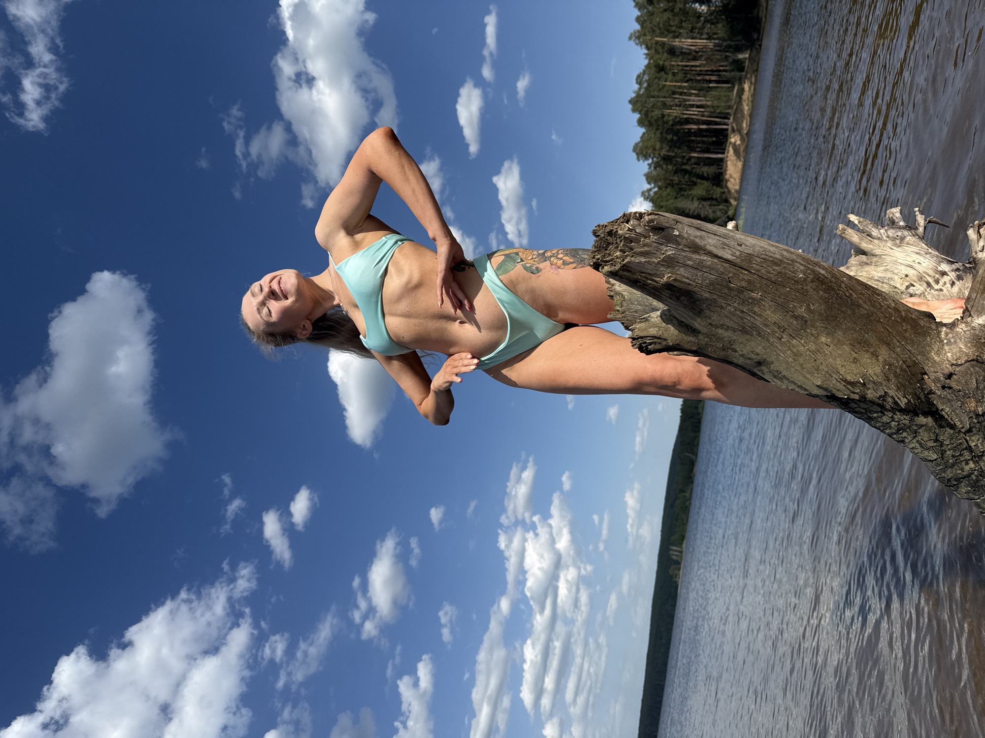 A woman in a light blue bikini stands on a dead tree trunk on the shore of a body of water. Her head is tilted back, and she is smiling with her eyes closed. She is standing with her left leg on the tree trunk and her right leg extended to the right. Her right hand is on her stomach and her left hand is raised. A tattoo is visible on her upper right thigh. The background consists of a blue sky with white clouds on the left, and water and trees on the right. The location is near the city of Chaikovsky, Russia.