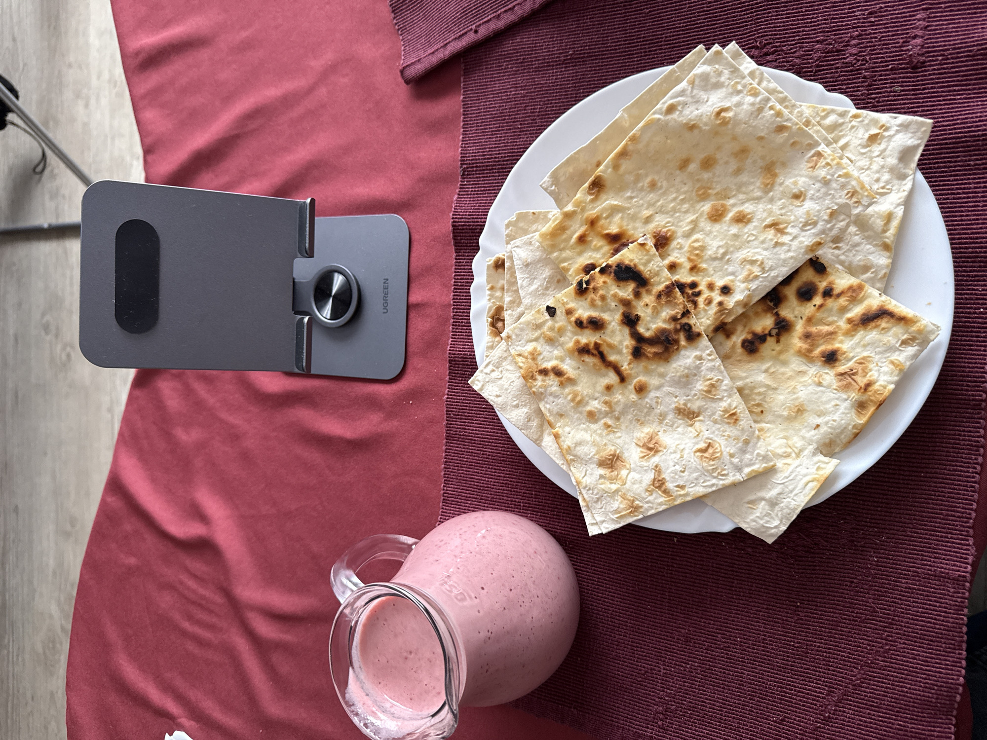 An overhead shot displays a table setup for a meal or snack, likely indoors during daylight hours, given the natural lighting. The scene is set on a red tablecloth, overlaid with a darker red, ribbed placemat that runs horizontally across the frame.
On the left side of the table, a dark gray metallic phone or tablet stand is visible, bearing the "UGREEN" brand name in white text. It features a black rubber grip and a circular, textured pivot point. To the right, resting on the placemat, is a white plate piled with several pieces of golden-brown flatbread, some showing distinct toasted or slightly charred marks.
In the bottom right corner, a clear glass pitcher or jug contains a thick, pink liquid, resembling a smoothie or milkshake. The background to the left reveals a light-colored wooden floor and a silver-toned metallic stand, possibly a tripod. The overall impression is a casual dining setting in Blagoevgrad, Bulgaria.