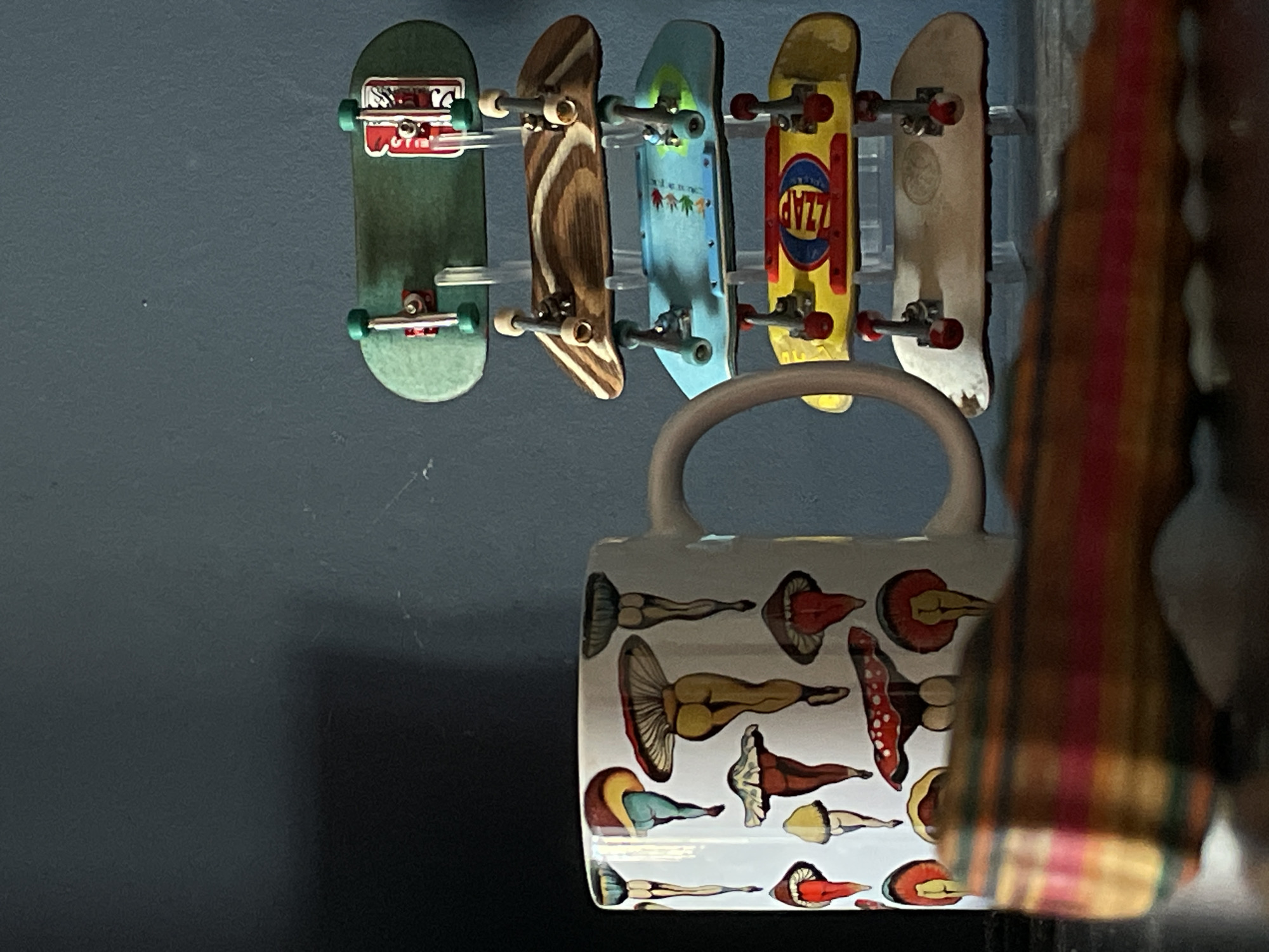 A medium shot shows five finger skateboards hanging on a wall above a ceramic mug decorated with mushroom images. The skateboards are arranged horizontally, facing the viewer. From left to right, they are colored green, brown, light blue, yellow, and white. The mug is positioned below the skateboards, partially obscuring the bottom of the skateboards. The handle of the mug is visible. A striped, blurred object stands in the foreground on the right side of the image, further obscuring the scene. The wall behind the skateboards is a muted blue-gray. The location is not discernable from the image.