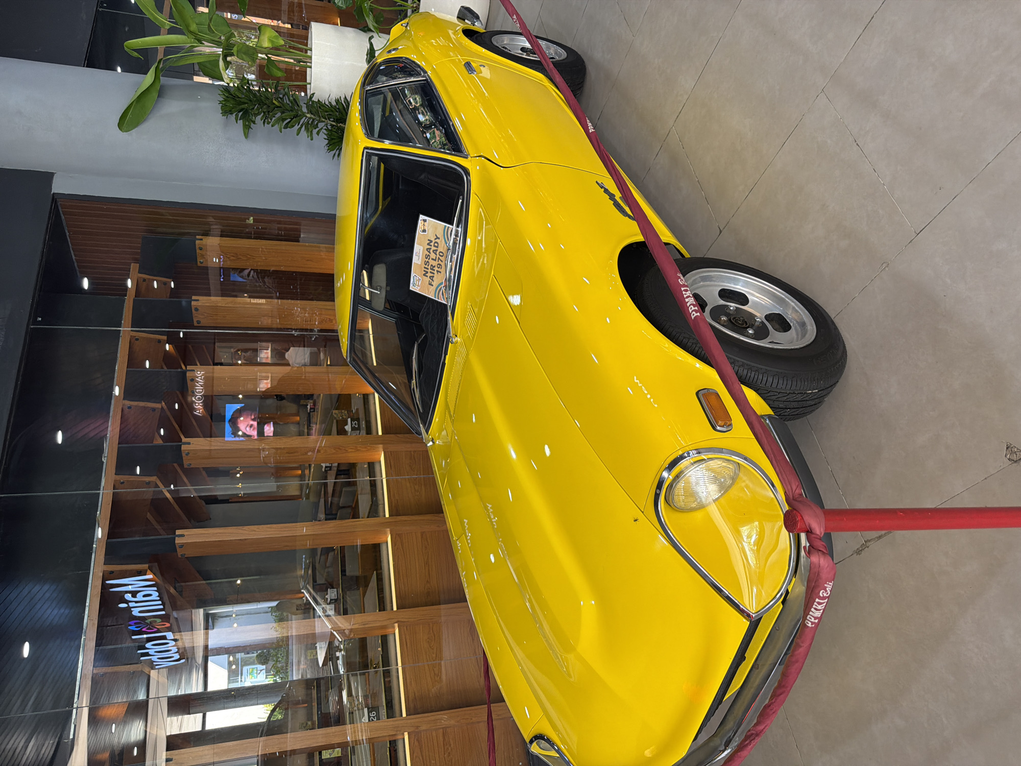 A vibrant yellow classic Nissan Fairlady 1970 automobile is prominently displayed indoors, likely in a commercial or exhibition space. The car is parked on a light grey tiled floor and is partially encircled by a red rope barrier wrapped around its front, bearing the white text "PPMKI Bali". An information sign on the car's windshield confirms its identity as a "NISSAN FAIRLADY 1970".
The setting appears to be a well-lit interior, possibly a main lobby, with reflections from artificial lighting visible on the car's polished surface. The background features modern architecture with large glass panels that reflect the surrounding environment. Visible through and in the reflections of the glass are wooden shelving or display units. Text reflected in the glass includes "Main Lobby" (reversed), "PANDORA" (reversed), and numbers "25" and "26". A screen inside the building, visible in one reflection, displays a person's face. Green potted plants are also present in the background, adding to the indoor decor. The scene suggests a static display of a classic vehicle in a public, commercial venue in Kuta, Indonesia.