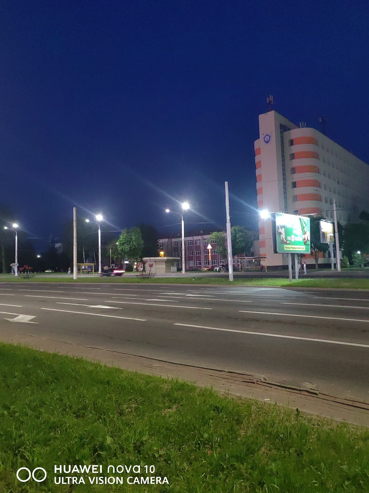 The image depicts an urban street scene at night in Vitsebsk, Belarus, captured by a Huawei nova 10 Ultra Vision Camera. The sky is dark blue, indicating late twilight or nighttime conditions, with artificial illumination being the primary light source.
In the midground-right, the distinctive Vitsebsk Regional Executive Committee building is prominently visible. This multi-story structure features light-colored walls (likely white or beige) adorned with multiple horizontal orange bands. Its roof hosts several antenna-like structures and HVAC units. A blue circular emblem, likely the coat of arms of the Vitsebsk Region, is affixed to the upper portion of the building's facade.
A multi-lane asphalt road occupies the midground and foreground, featuring clear white lane markings, including two solid white lines and a white arrow indicating a forward or turning direction. Green grass covers the foreground and the median separating the road lanes.
Along the road, numerous tall streetlights emit bright white light, casting distinct lens flares. On the right side of the road, in front of the executive committee building, a large, illuminated billboard displays an advertisement with a green background and Cyrillic text, reading "НА" and "Найдай тавары на свой густ" (Find goods to your taste). A smaller, partially visible billboard is situated behind it. A light-colored, possibly concrete, bus shelter is located nearby.
On the left side of the road, farther down, several older-style multi-story buildings with illuminated windows are visible. A yellow and white bus shelter is positioned in the midground-left, near a prominent tree with bright green foliage. Overhead trolleybus wires are faintly visible crisscrossing the sky above the road.
Several individuals are present on the sidewalks and near the bus stops, discernible as indistinct figures due to distance and lighting. A dark-colored car with blurred taillights is in motion on the road in the midground-center, while a lighter-colored car is parked or slowly moving on the far left. Lush green grass covers the foreground, with a visible concrete curb and a sliver of sidewalk bordering the road.