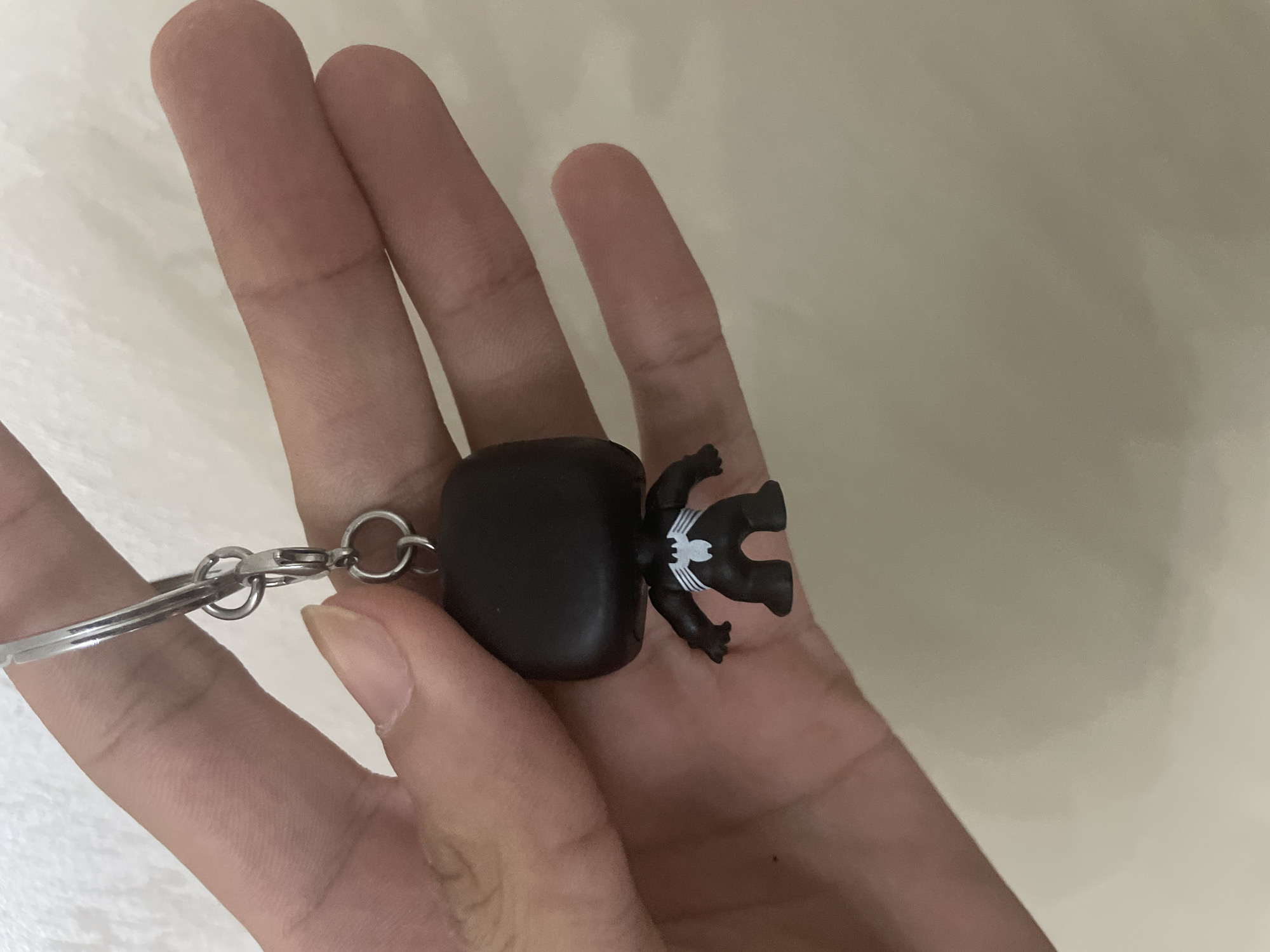 A human hand, with light skin tone and visible knuckles and fingernails, is centrally positioned, holding a silver-colored metallic keychain. The keychain consists of a main ring, a short chain, a lobster clasp, and an additional small ring from which a miniature figurine is suspended. The figurine is a stylized, black, squat character with a large head and small body, consistent with a Funko Pop! design. A prominent white spider emblem with elongated legs is visible on the character's upper back. The hand's fingers are slightly curled, gripping the metal rings of the keychain. The background is a plain, light, off-white or beige wall, lacking any distinguishing features or discernible depth. There is no visual information within the image to indicate the location as Ihtiman, Bulgaria.