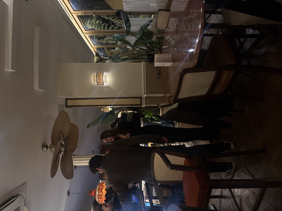The image captures an interior view of a restaurant or bar located at 20 Rue du Roi Doré in Paris, France. The scene is dimly lit with warm artificial light, featuring several individuals and characteristic furnishings.
In the left foreground, a man with dark hair, wearing a dark long-sleeved top, stands with his back partially to the viewer, positioned near a bar counter. To his right, a woman with long dark hair, dressed in a dark top and pants, stands facing slightly right, engaged in conversation or observation. Other indistinct figures are visible further back near the bar.
The bar area on the left features a counter illuminated by a blue under-light, displaying bottles, glasses, and a tablet device. An orange-red, scalloped pendant light fixture hangs above this section. Overhead, a large, ornate ceiling fan with multiple woven blades is mounted on a white ceiling, which also includes an air conditioning vent.
The right side of the frame showcases a polished, dark wooden oval dining table reflecting ambient light. It is surrounded by chairs with dark wooden frames, cane backs, and brown upholstered seats. A menu or brochure stands upright on the table. A dark jacket or coat is draped over the back of one chair in the foreground. The walls are light beige with a darker beige wainscoting and decorative vertical panels featuring a cane weave pattern. Wall-mounted sconces with woven shades emit warm light.
Large windows along the right wall reveal dense green foliage, including ferns and large-leafed plants, situated directly outside or in a heavily planted interior section. The floor throughout the establishment is composed of dark wood laid in a herringbone pattern. Several large potted tropical-style plants are strategically placed, contributing to the establishment's decor.