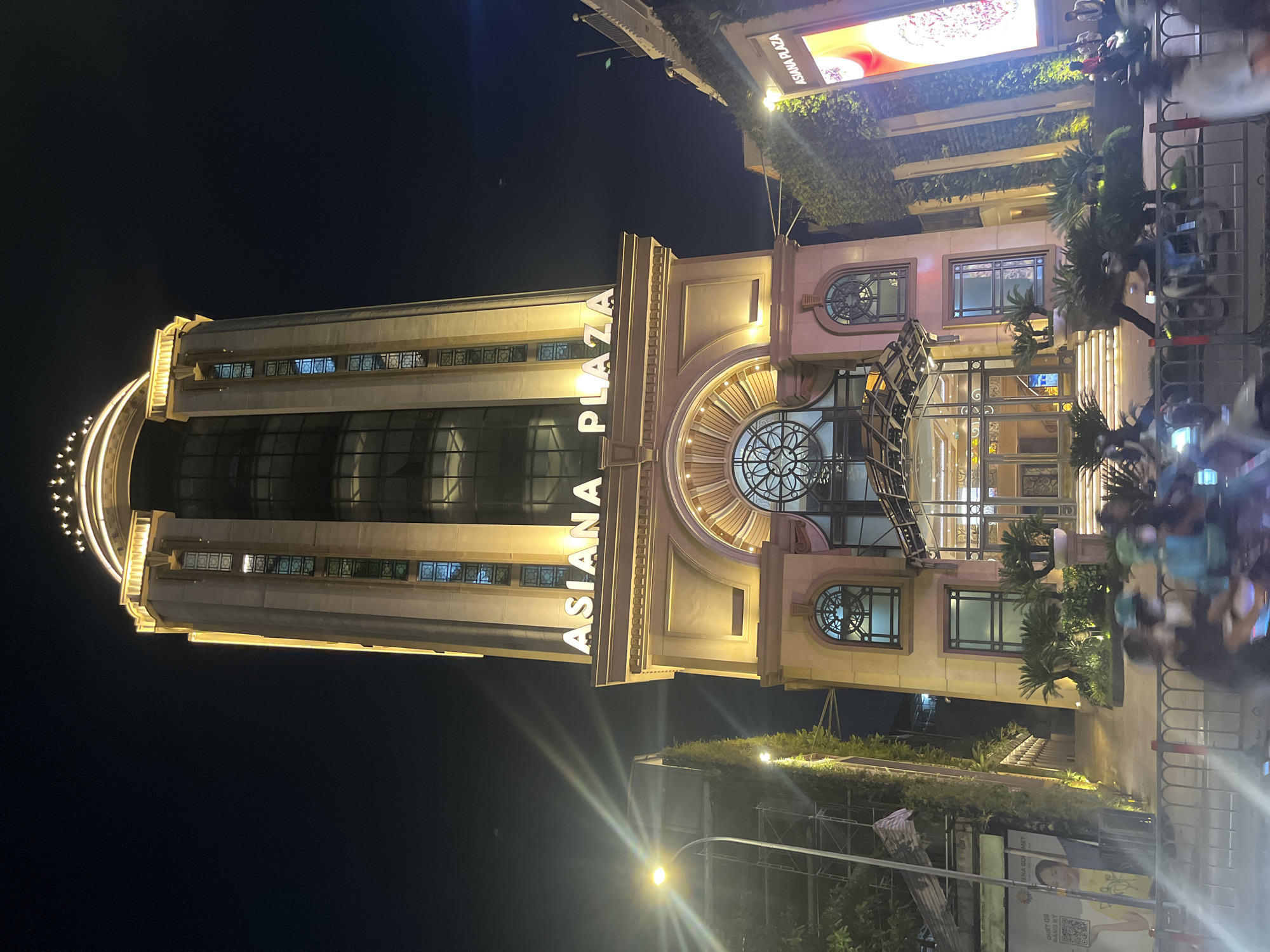 This is a low-angle, medium shot of the ASANA PLAZA building at night, located in an urban setting. The building is a tall, multi-story structure with illuminated signage reading "ASANA PLAZA" in large, white, capitalized letters. The facade features a mix of concrete, glass, and decorative architectural elements, with warm yellow lighting accentuating its features. At the top, the building is crowned with a decorative illuminated element resembling a tiered crown.
The entrance to the building is grand, with a large, arched, glass-enclosed entryway featuring an ornate circular design in the glass above. Steps lead up to the entrance, and there are palm plants and other greenery flanking the sides.
To the left of the ASANA PLAZA building, another building is visible, partially obscured by scaffolding and covered in dense green foliage. A streetlamp with a bright light is positioned on the left side of the frame, casting beams across the scene.
The foreground shows a street with several motorcycles and people blurred by motion, suggesting they are in transit. A metal barrier with red and white markings runs along the bottom of the frame, separating the street from the sidewalk or building entrance area. A large illuminated billboard is visible on the right side of the ASANA PLAZA building, displaying the "ASANA PLAZA" logo and an image. The sky above is dark, indicating nighttime. The overall scene conveys a busy urban environment at night, with the ASANA PLAZA building being the central illuminated subject.