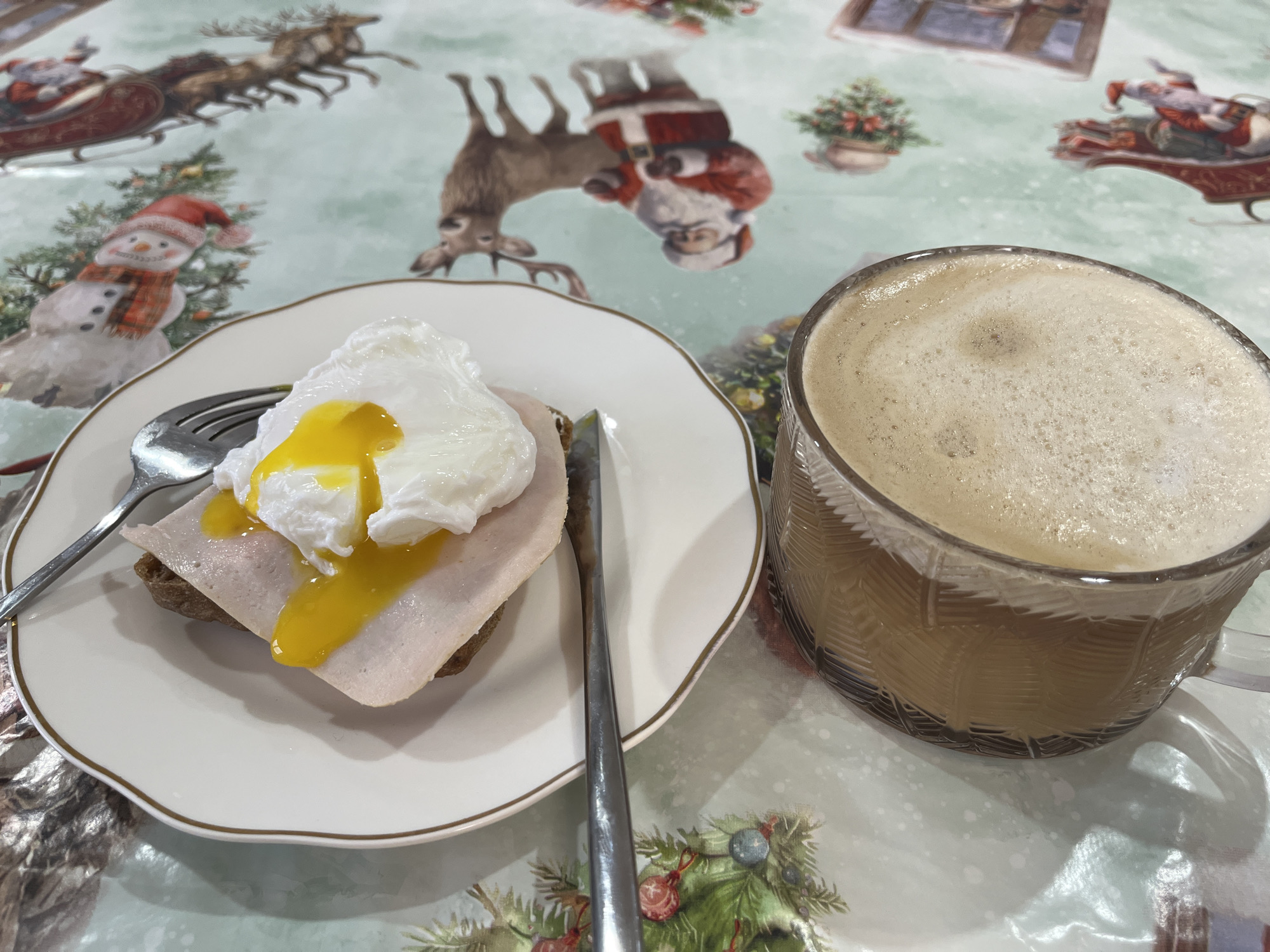 This close-up image presents a breakfast or brunch scene, featuring a plate of food and a beverage set on a festive, Christmas-themed tablecloth. On a white plate with a gold rim, a slice of dark bread or toast is topped with several layers of light-colored deli meat, possibly ham or turkey. Perched on top is a perfectly poached egg, with its firm white surrounding a vibrant, runny yellow yolk that spills slightly over the meat. A silver fork rests on the left side of the plate, while a silver knife lies to the right.
Beside the plate, a clear glass mug is filled with a frothy, light brown beverage, likely coffee or a latte, indicating a morning or early afternoon meal. The background is dominated by the tablecloth, which features a cheerful, light green design adorned with various Christmas illustrations. Visible motifs include Santa Claus riding in a sleigh pulled by reindeer, a friendly snowman wearing a red hat and scarf beside a decorated Christmas tree, and other festive holiday elements. The overall setting evokes a cozy and celebratory atmosphere, characteristic of the holiday season. The location context for this image is Chaikovsky, Russia. No visible text is present in the media file.