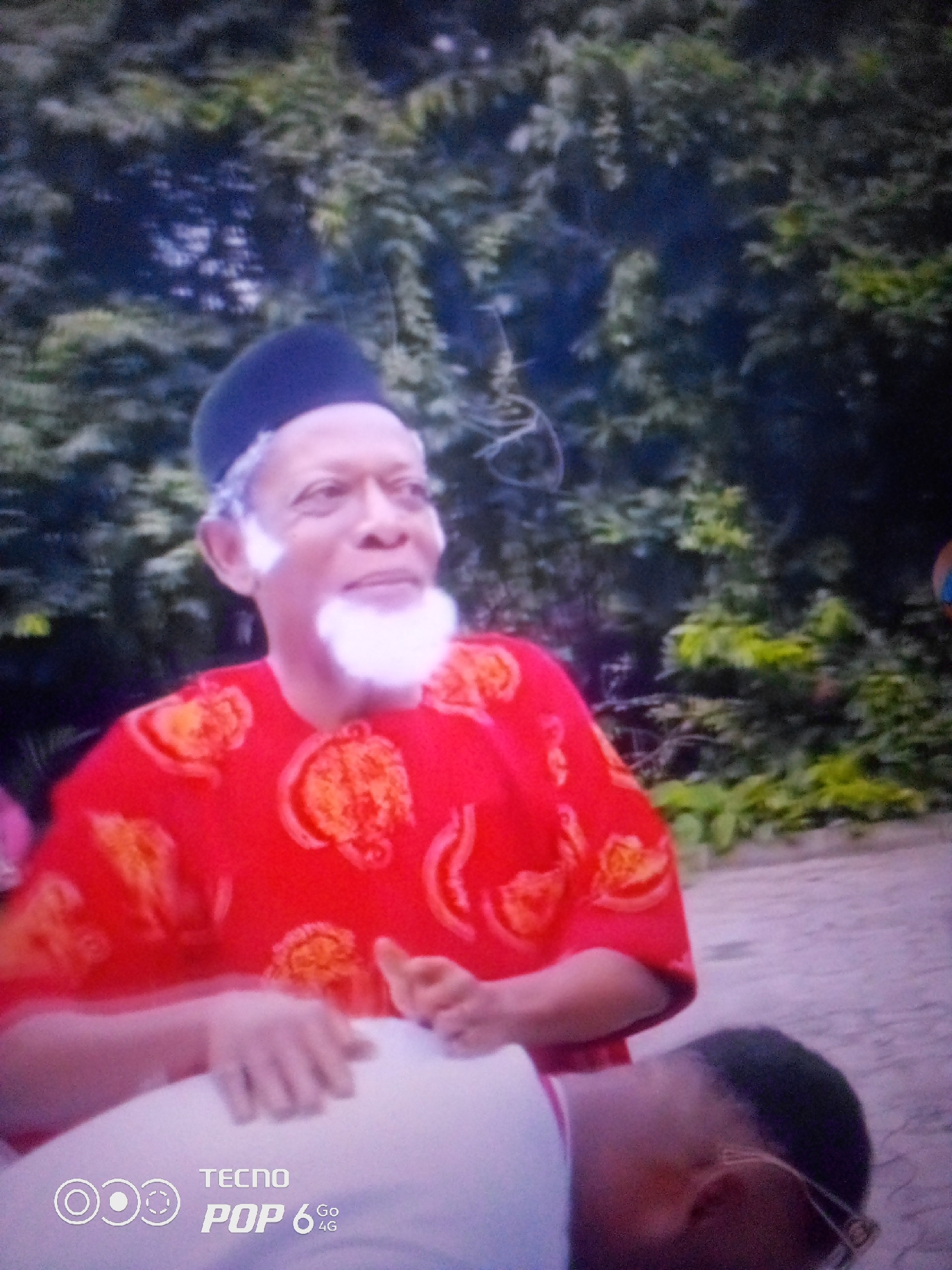 An older man wearing a dark kufi and a red shirt with an orange pattern is pictured outdoors. He has a white beard and mustache. His eyes are looking to the right. He is wearing a white shirt below the red shirt. Another person's head and shoulders are visible in the foreground, with their face turned away from the camera. The background consists of dense green foliage, suggesting an outdoor setting possibly in a rural or park area. The image also has a watermark in the bottom left corner that reads "TECNO POP 6 Go 4G." The scene does not provide any specific indicators of the city of Howayi, Nigeria.
