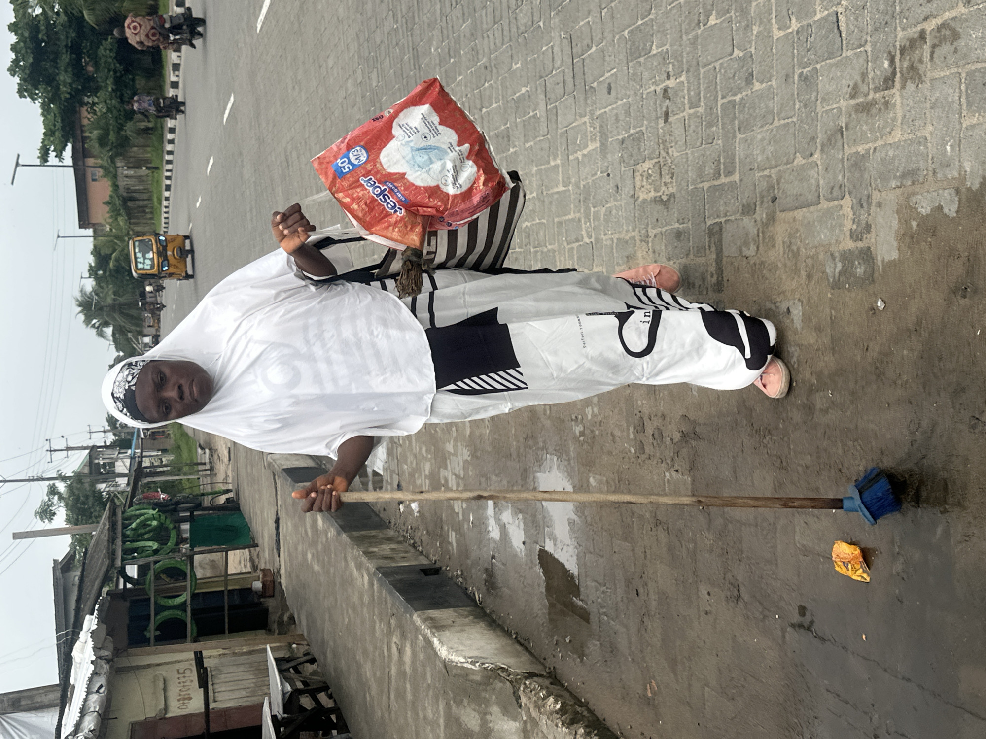 A woman wearing a white hijab and a white and black patterned dress walks on a paved street in Badagry, Nigeria, carrying a broom and two shopping bags. The street is wet, with puddles visible. On the left side of the street, there are several small shops with corrugated metal roofs. In the background, there is a road with several vehicles, including a yellow auto-rickshaw, and two motorcycles. The sky is overcast. The woman appears to be looking directly at the camera with a neutral expression. One of the shopping bags is red and contains baby diapers. The other bag is a striped tote bag. A piece of orange trash is on the ground near the broom.