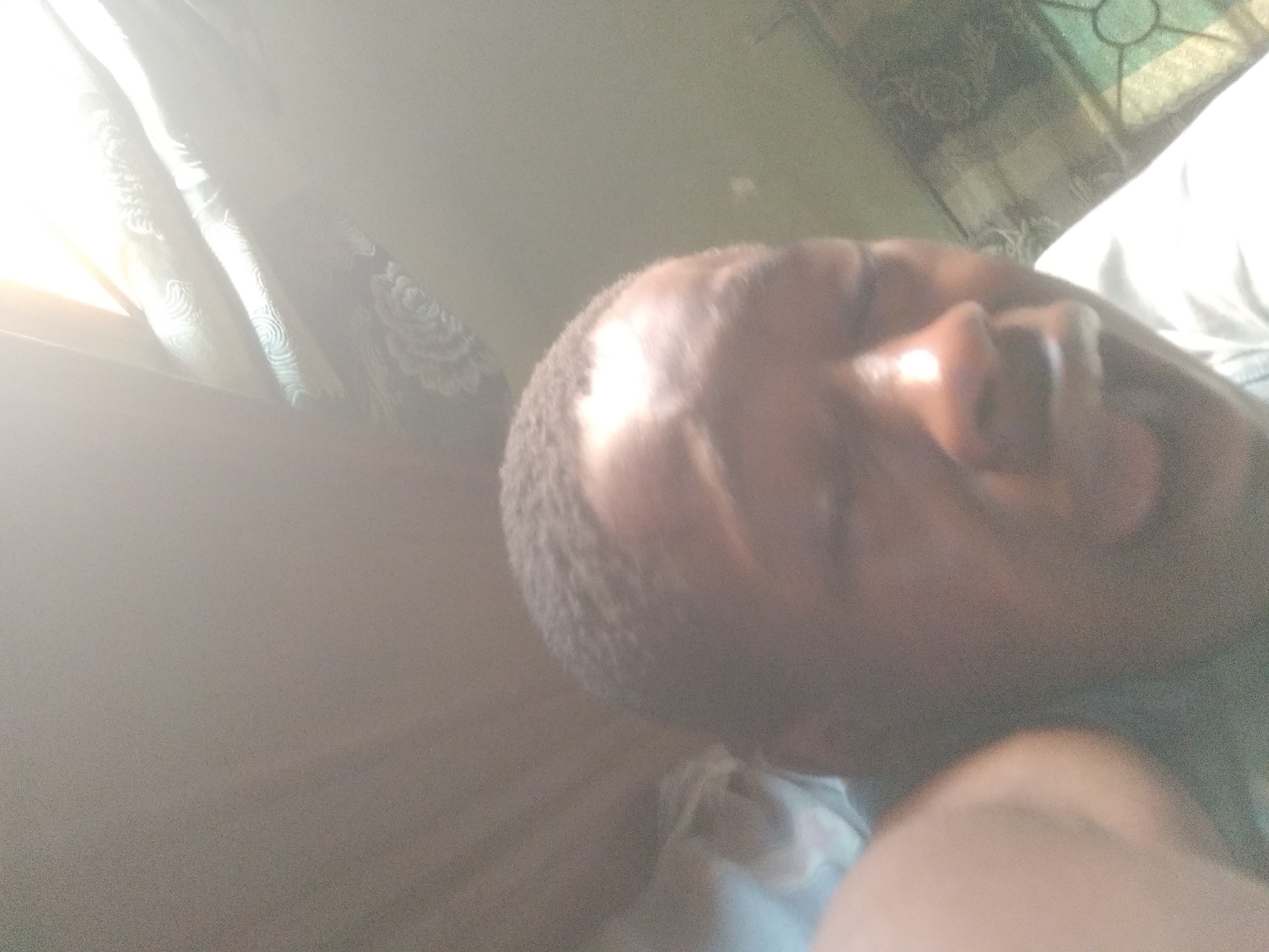 The image appears to be taken indoors. An individual with short, dark hair is lying down. The person's face is angled toward the camera. A window with patterned curtains is visible in the background. Light is streaming in through the window. The location appears to be Shangisha, Nigeria.