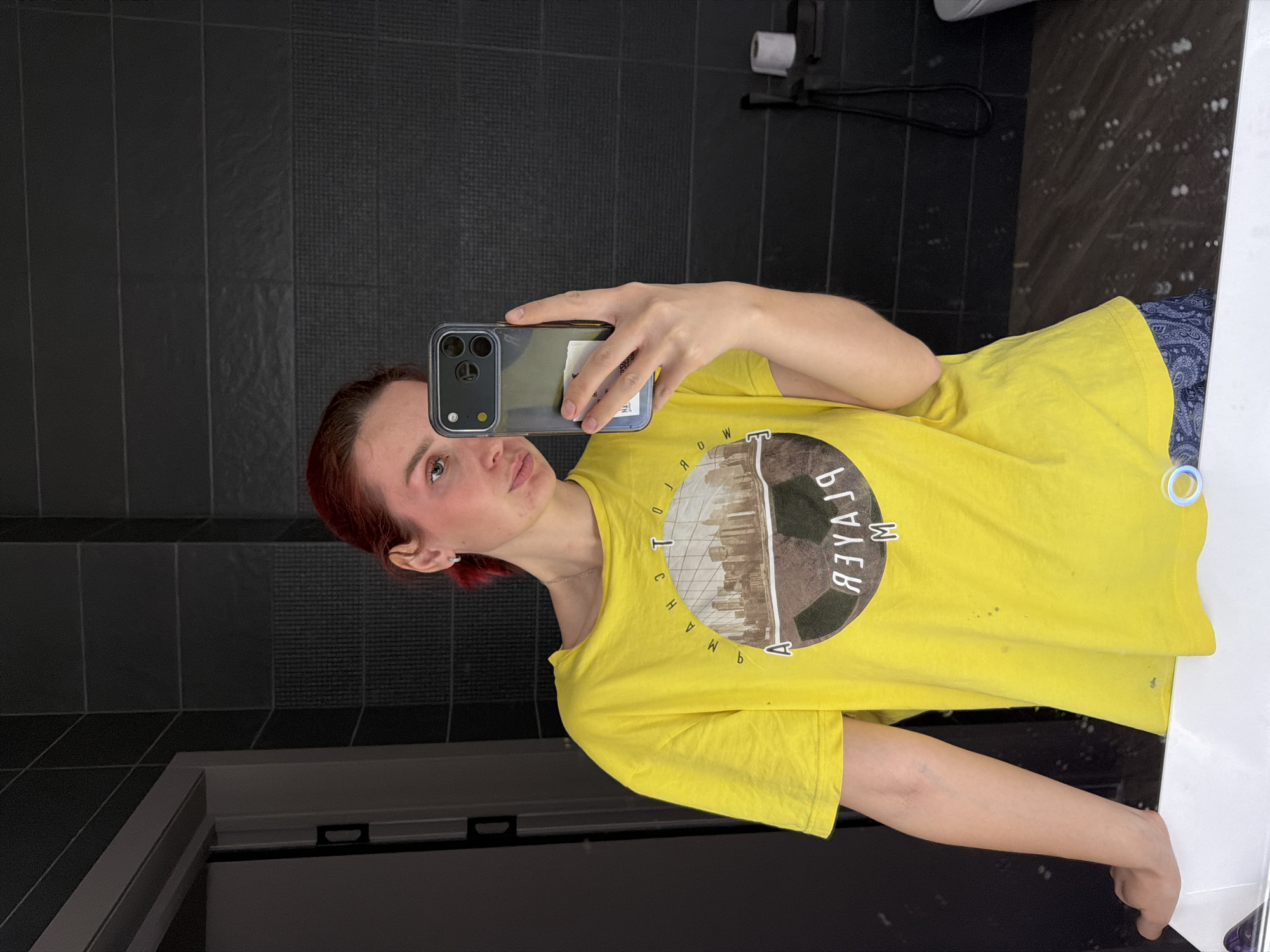 A woman takes a selfie in a bathroom. The woman is fair-skinned with short, reddish-brown hair. She is wearing a yellow t-shirt with a black and gray graphic design on the front. She holds a smartphone with a protective case up to her face, partially obscuring her features. The background consists of dark gray, vertically-oriented rectangular tiles. A toilet paper roll is visible on a black fixture mounted to the wall. Below the woman, a white countertop and a dark gray floor can be seen. The bathroom could be located in the city of Chaikovsky, Russia, if that is the person's location.