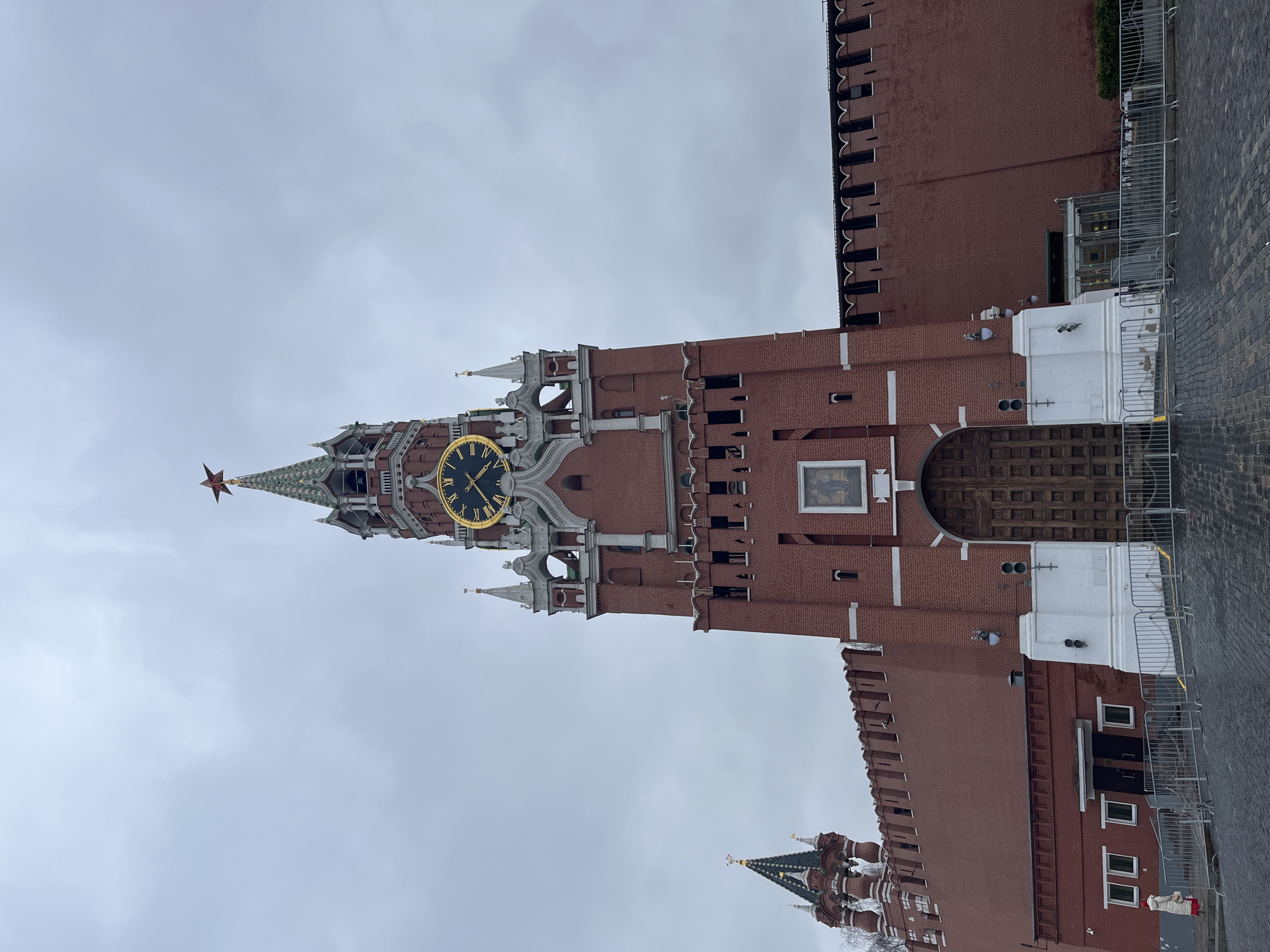 The image shows the Spasskaya Tower of the Moscow Kremlin in Moscow, Russia. The tower is constructed of red brick and features a large clock face, a red star on top, and various architectural details. Part of the Kremlin wall is also visible, along with barriers at the base of the wall. The sky is overcast.