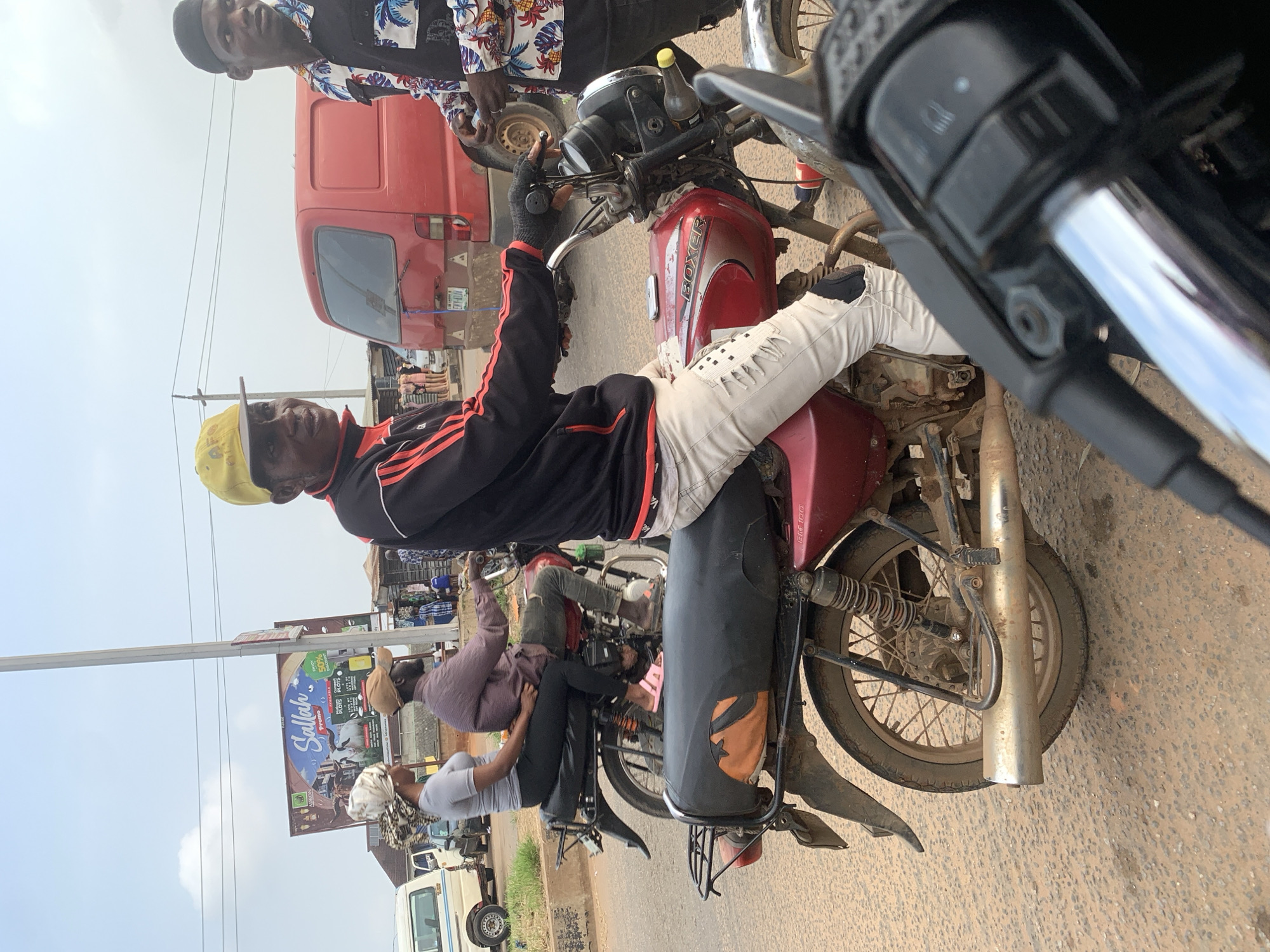 This image captures a street scene in Shagamu, Nigeria, with a focus on motorbikes and their riders. In the foreground, a person wearing a yellow cap and a black and red jacket is seated on a red motorcycle, with their right hand on the handlebars. This individual is looking towards the left of the frame. Behind them, another person in a colorful shirt is standing and looking to the right. Further back, two people are riding on a separate motorbike, with the driver wearing a brown cap and the passenger in a grey top and headscarf. The background shows a red van, a white vehicle, a billboard for "Sallah Promo," and various market stalls. The ground is dirt and dusty, indicative of a road in a developing urban area. The sky is overcast.