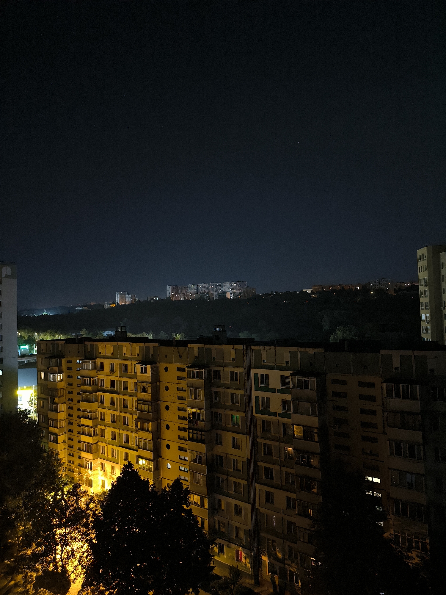 The image is a night-time, high-angle photograph of a residential urban landscape in Chișinău, Moldova. In the foreground, a large, multi-story apartment building with a light yellow facade and numerous balconies is prominently featured. The building is illuminated by artificial lights from within, with many windows showing a warm, yellow glow. Trees with dark, silhouetted foliage are visible in front of the building, partially obscuring its lower levels.
Further in the background, across a hilly, tree-covered expanse, are more apartment buildings of varying sizes and architectural styles, also lit from within. These buildings are spread across the elevated terrain, with some appearing to be older Soviet-era structures and others possibly more modern. The sky above is a deep, dark blue, with a few faint stars visible. The overall impression is one of a sprawling urban neighborhood at night, with the glowing windows of residential buildings creating a sense of life and activity amidst the darkness.