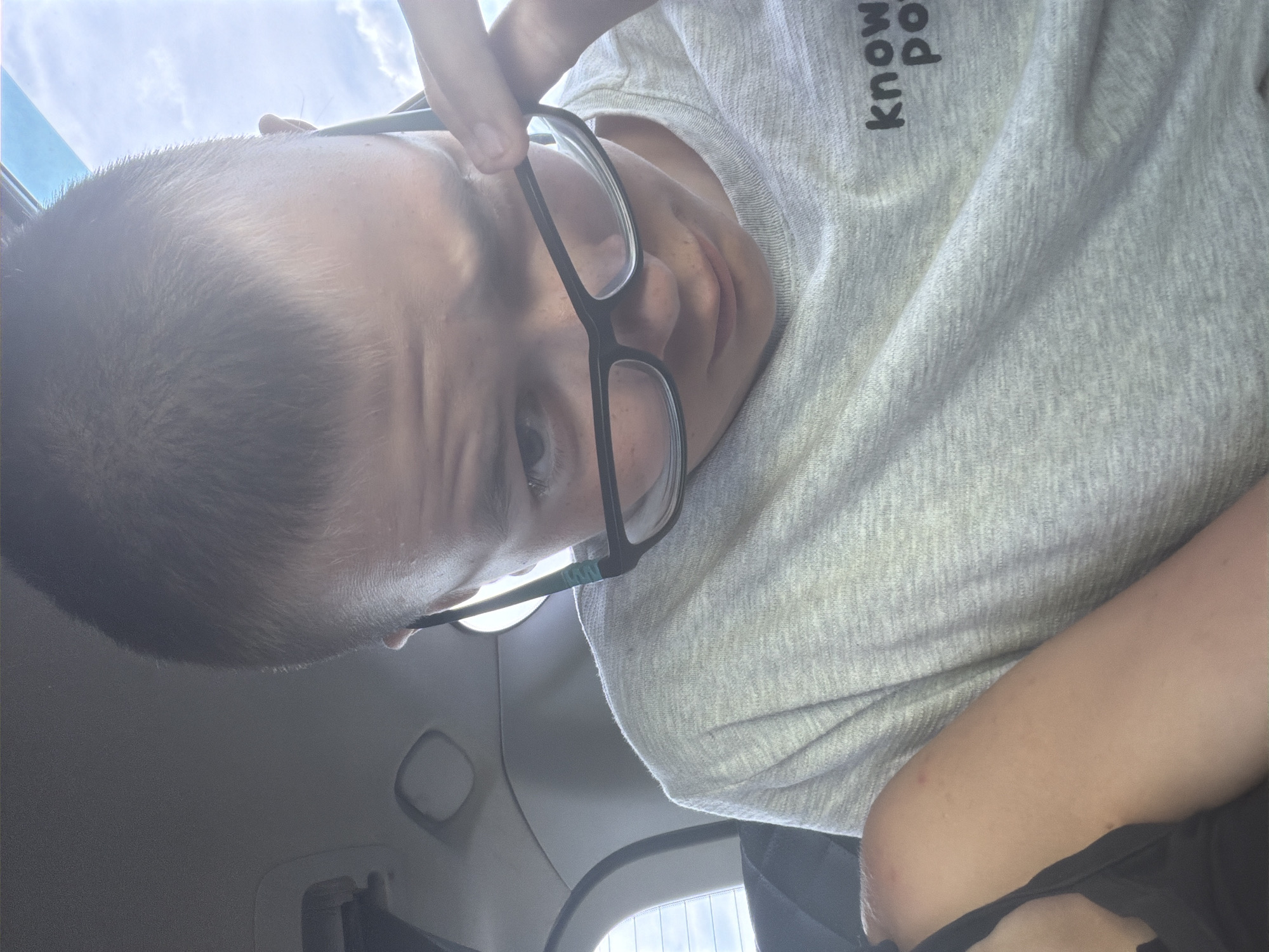A young person with short brown hair, wearing glasses and a light gray t-shirt with the text "know po" printed on it, is captured in a close-up, selfie-style photograph. The person is looking down and to the side, with their right hand resting on their glasses. They appear to be inside a vehicle, indicated by the presence of car interior elements such as a headrest and a window with a visible sky and clouds. A portion of another person's arm, visible on the left side of the frame, suggests the photographer is not alone. The lighting is bright, possibly indicating daylight. The background shows the interior of a car, with the sky visible through the windows. The photograph is likely taken in Teneevo, Russia, based on the provided context.