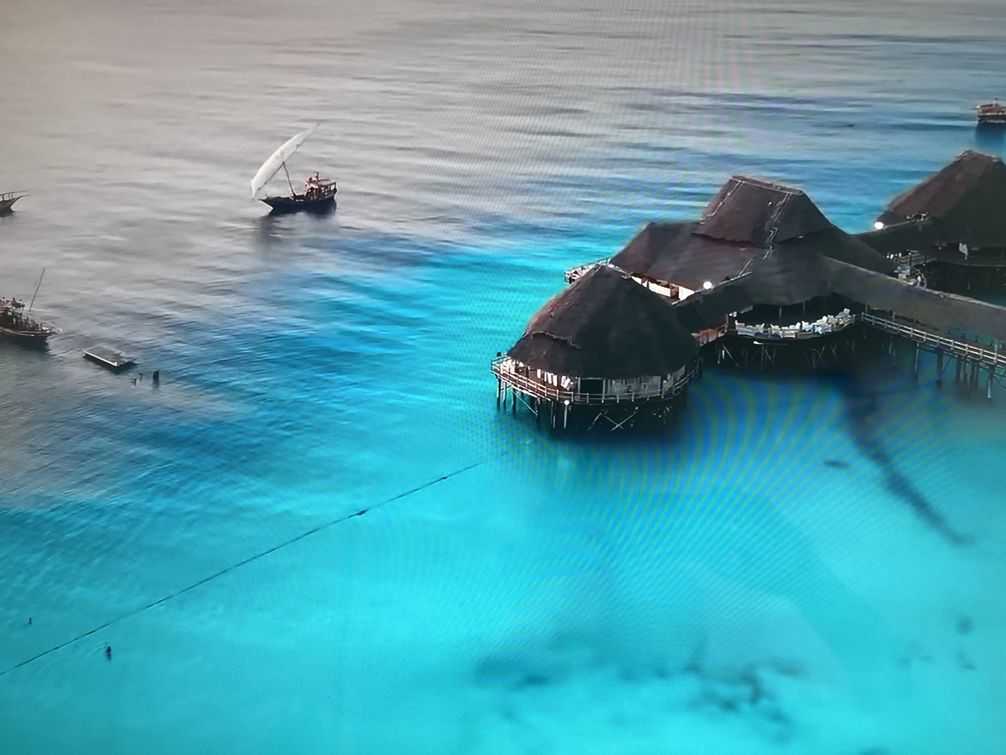 The image shows an aerial view of a tropical coastal scene, likely a resort area with buildings on stilts over clear, turquoise water. A group of thatched-roof buildings is connected by a wooden pier. Several boats are visible on the water, including one sailboat. The water clarity suggests a shallow, sandy bottom. Two individuals can be seen swimming. The scene does not appear to be in Basingstoke and Deane, United Kingdom.
