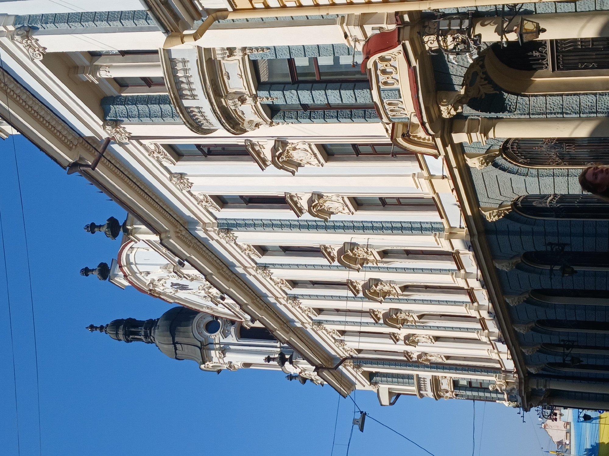 The image is a low-angle, medium shot of the ornate facade of a building in an urban environment, under a clear blue sky. The building features intricate architectural details, including columns, decorative moldings, arched windows, and balconies. The facade is painted in a light cream color with blue accents on the lower levels and around some window frames. A dark dome-topped tower with decorative spires is visible on the left side of the building. Power lines and a street lamp are also visible in the foreground. In the lower right corner, a person's face is partially visible, looking slightly to the left. The street level features arched entrances with decorative grates.