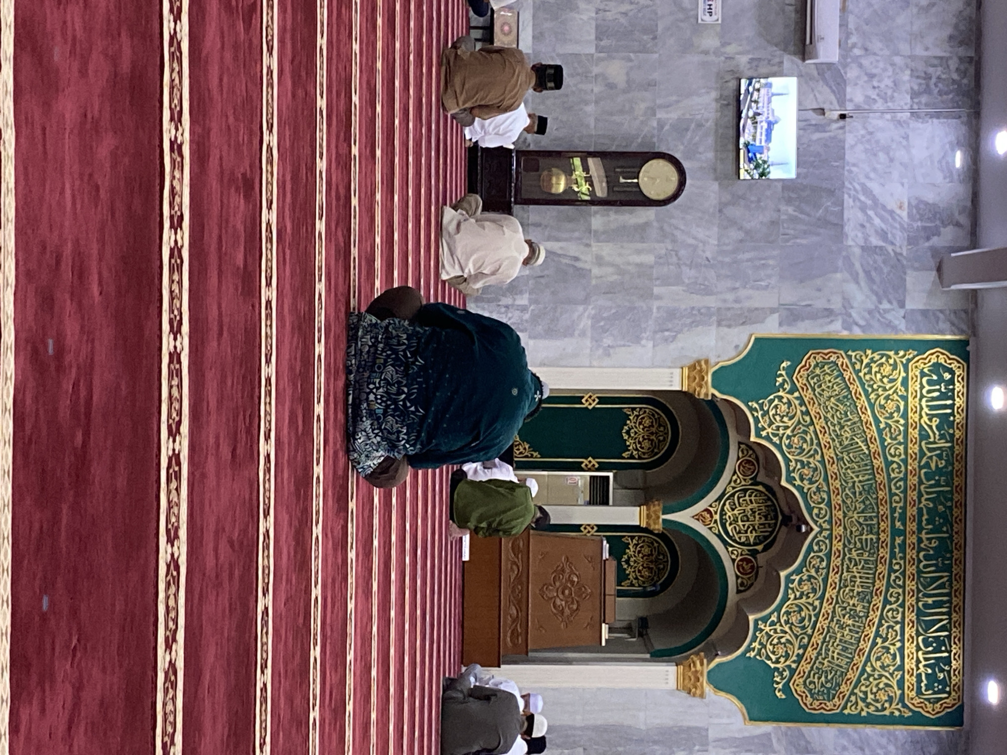 The image depicts the interior of a mosque, specifically the Baiturrahman Grand Mosque in Lhokseumawe, Indonesia. Several men are seated on a red carpet with striped patterns, facing away from the camera. The men are engaged in prayer or contemplation. The mosque's interior features elegant green and gold Islamic calligraphy on the wall behind the men. A large wooden podium is positioned in front of the calligraphy. The walls are made of marble, and a large grandfather clock stands to the left of the podium. A television screen displaying a mosque building is mounted on the left wall, above the clock. The lighting appears to be artificial.