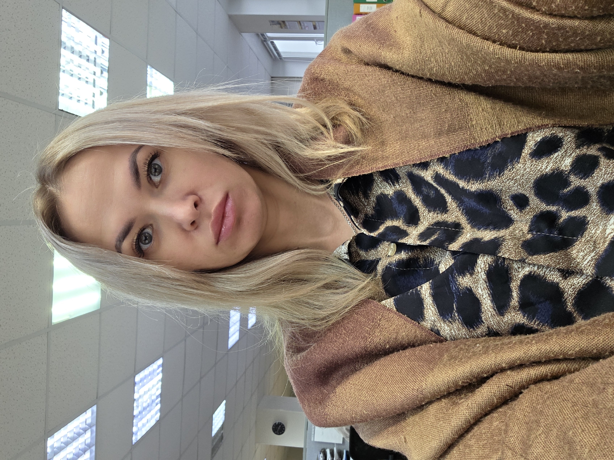 A woman with light blonde hair and light-colored eyes is posing for a selfie. She is wearing a leopard-print top under a brown shawl or wrap. The background appears to be an office setting with fluorescent lighting panels in the ceiling. The location is Chaikovsky, Russia.