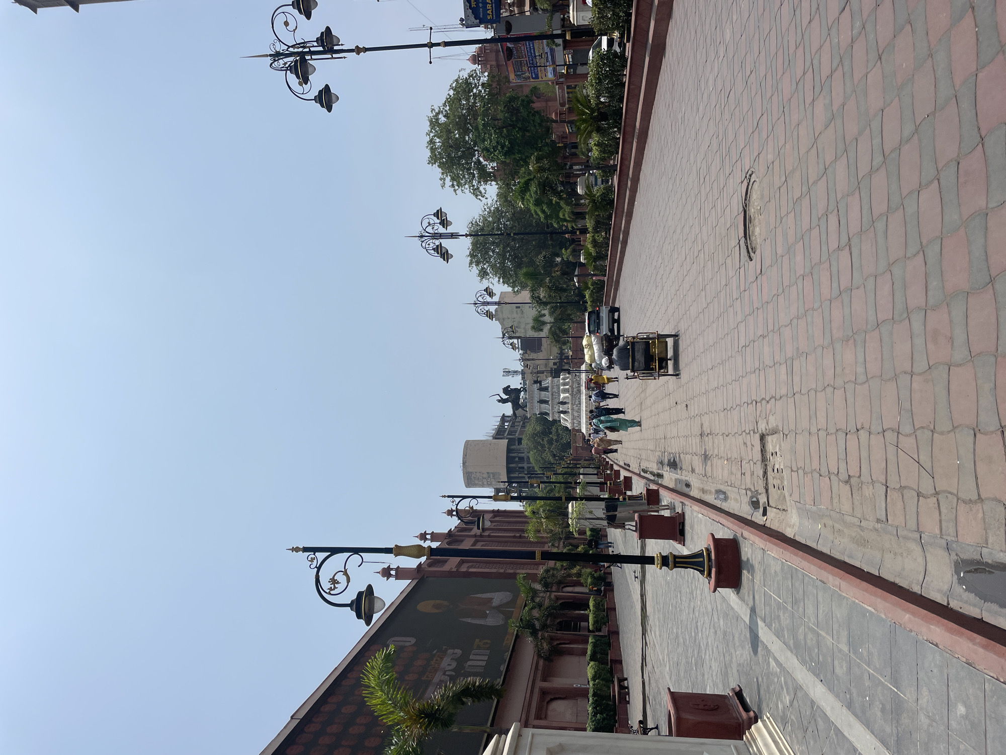 The image shows a street scene in Amritsar, India, likely in the vicinity of the Golden Temple, given the style of architecture and the presence of people in traditional attire. The street is paved with red and grey interlocking bricks, and bordered by a red curb. On the left, a large building with a red facade and ornate windows is visible, featuring a large digital screen displaying an image of a man. Several palm trees and other greenery line the sidewalk. Ornate black lampposts with gold accents are placed along the street.
In the distance, a monument featuring statues of horses and a rider stands in the center of a roundabout. People are walking along the sidewalks and crossing the street. A few vehicles, including a white car and a small cart pulled by a person, are present on the road. The sky is clear and blue. The overall impression is a busy, well-maintained urban environment with a clear cultural and historical significance.