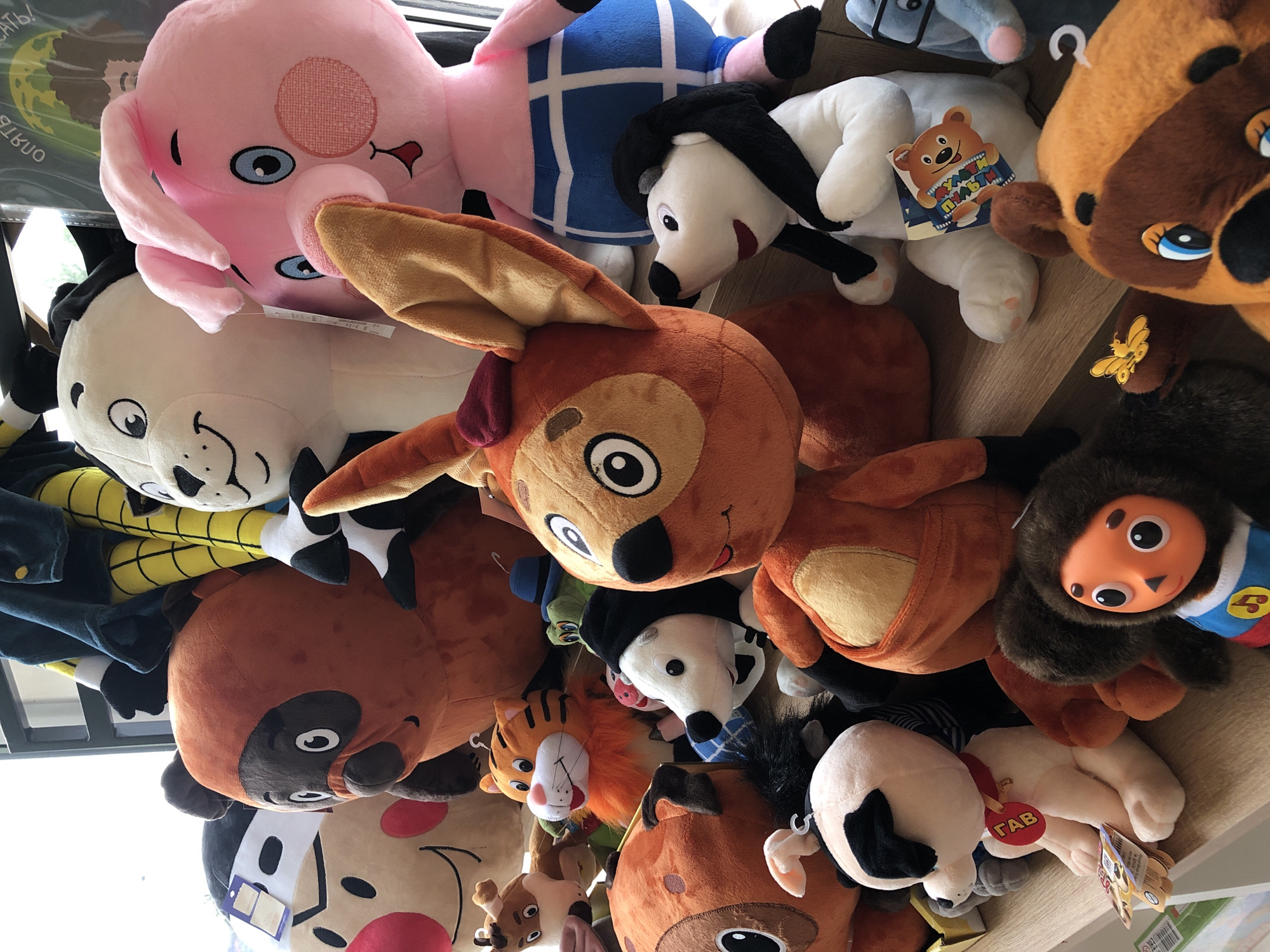 A collection of stuffed animals is displayed on wooden shelves, presumably in a retail setting, given the presence of tags and packaging. The stuffed animals vary in size and character, including a large pink pig, a brown bear-like character, a white dog with black ears, a tiger, and other animal figures. Some are stacked, while others are placed individually. The lighting suggests an indoor environment, possibly a shop or a home display. The overall scene is static, with no human activity depicted.