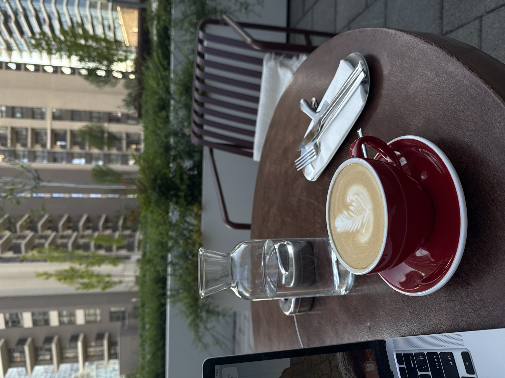 The image displays an outdoor café or patio scene, focusing on a dark brown, textured round table. On the table, a vibrant red ceramic cup and saucer hold a latte with a prominent rosetta-style latte art pattern. Adjacent to the coffee, a clear glass carafe or bottle contains water. Further back, a silver fork and knife are neatly placed on a white napkin, which rests on a small silver tray.
In the bottom right corner, a portion of a silver laptop, likely a MacBook, is visible, showing keys such as "delete," "return," and "shift" on its keyboard. A digital clock on the laptop screen reads "18:23," indicating the time as 6:23 PM.
The background reveals a brown slatted outdoor chair. Beyond the patio furniture, several modern multi-story buildings with structured balconies and glass facades rise into the urban skyline. Green foliage and trees are interspersed throughout the background, adding natural elements to the concrete environment. The scene suggests a calm and relaxed individual activity, possibly working or enjoying a coffee break in an urban setting. The provided context indicates the location is Belgrade, Serbia. No people are visible.