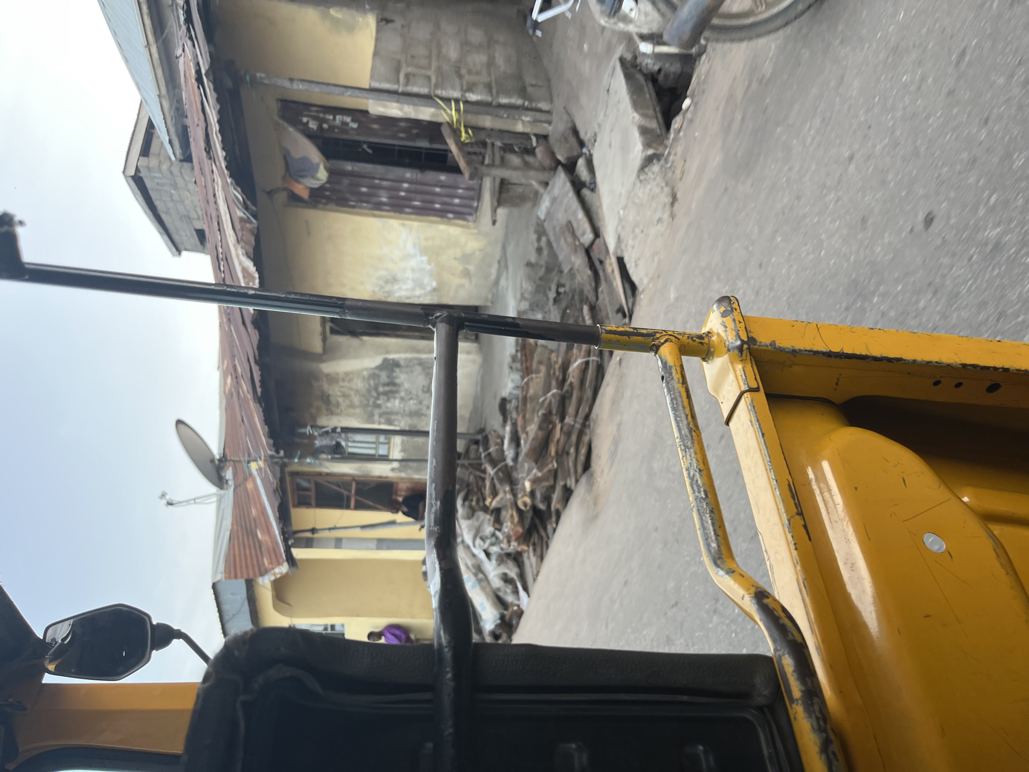 The image, taken from the perspective of a passenger in a yellow vehicle, captures a street scene in an urban environment. In the foreground, the yellow vehicle's frame and a rearview mirror are visible. The background reveals a street lined with buildings. Some buildings have corrugated metal roofs and appear weathered, with peeling paint and visible brickwork. In front of the buildings, a pile of chopped wood or logs is stacked on the ground. A few people are present in the scene: one individual in a purple garment stands near a building, and another person is partially visible indoors. The street itself is paved, and a satellite dish is mounted on one of the rooftops. A motorcycle is parked on the side of the road. The overall impression is one of a developing or informal urban setting.