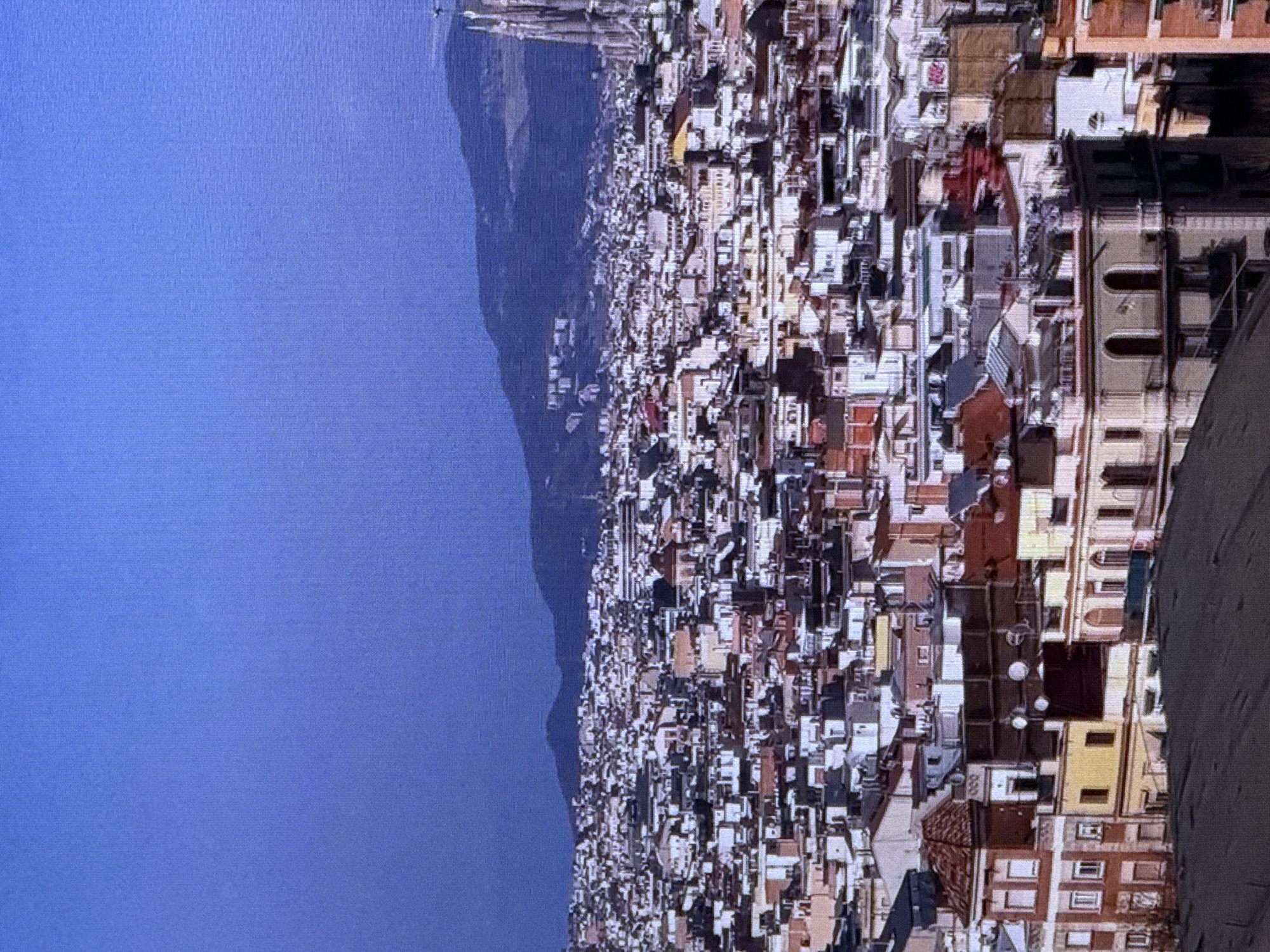 The image shows a panoramic view of a densely packed cityscape with numerous buildings. The architecture appears to be a mix of residential and possibly commercial structures, characterized by various roof styles, window placements, and building heights. Many of the buildings have tiled roofs in shades of red and brown, with facades in white, yellow, and muted earthy tones. Some buildings feature balconies and decorative elements. In the background, a mountain range can be seen under a clear, deep blue sky. To the far right, a tall, Gothic-style spire or tower is visible, suggesting the presence of a significant landmark, possibly a church or cathedral. The overall impression is of an established urban area.
There is no indication in the image that this scene is located in Dubai, United Arab Emirates. The architecture and landscape are not consistent with typical Dubai urban environments, which are characterized by modern skyscrapers and distinct desert aesthetics.