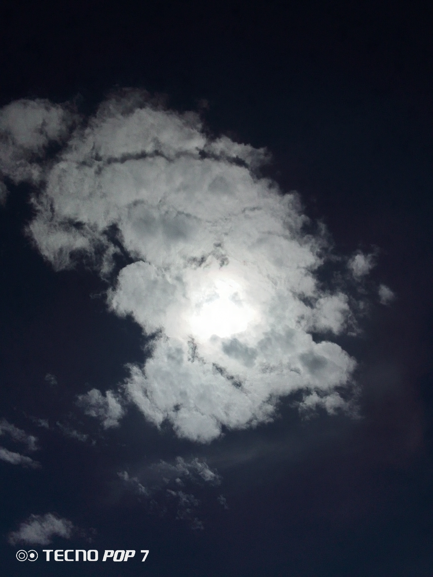 The image captures a view of the sky, with a large, bright cloud partially obscuring the sun. The sky is a deep, dark blue, almost black, providing a stark contrast to the white and gray of the cloud formation. The sun's light shines through the center of the cloud, creating a luminous effect. The photo was taken with a TECNO POP 7 phone. The city of Pedro González, Venezuela is not discernible from this image.