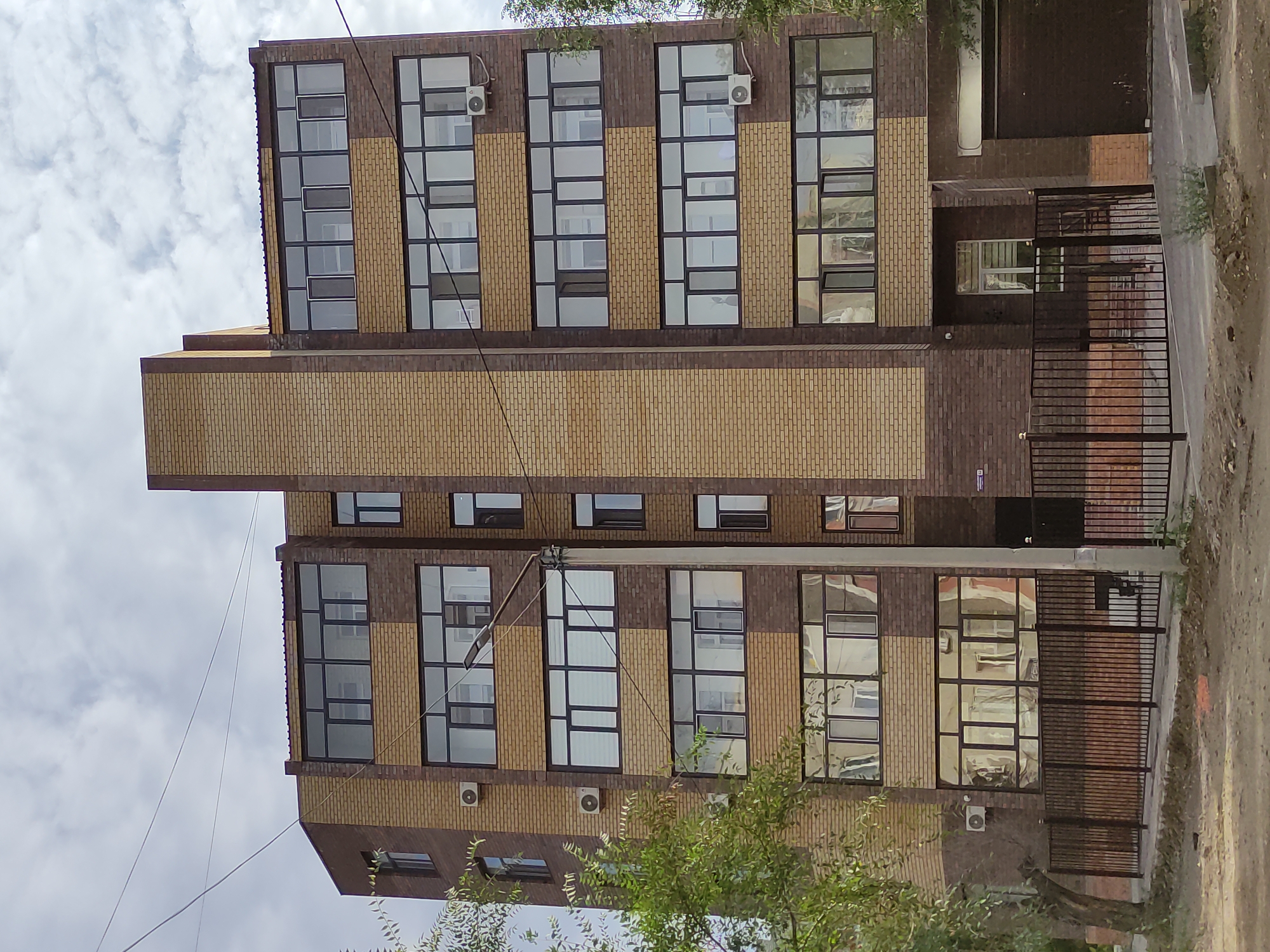 The image shows a modern, multi-story brick building in the city of Semey, Kazakhstan. The building features a combination of light yellow and dark brown brickwork, with large, dark-framed windows on each level. Several air conditioning units are visible on the exterior walls. A tall, central brick pillar runs vertically up the facade. The building is partially obscured by green trees on the left side. A dark metal fence runs along the front of the property, with a dirt road or construction area in the foreground. A street lamp is positioned near the center of the building. The sky above is cloudy. The building appears to be residential or commercial.