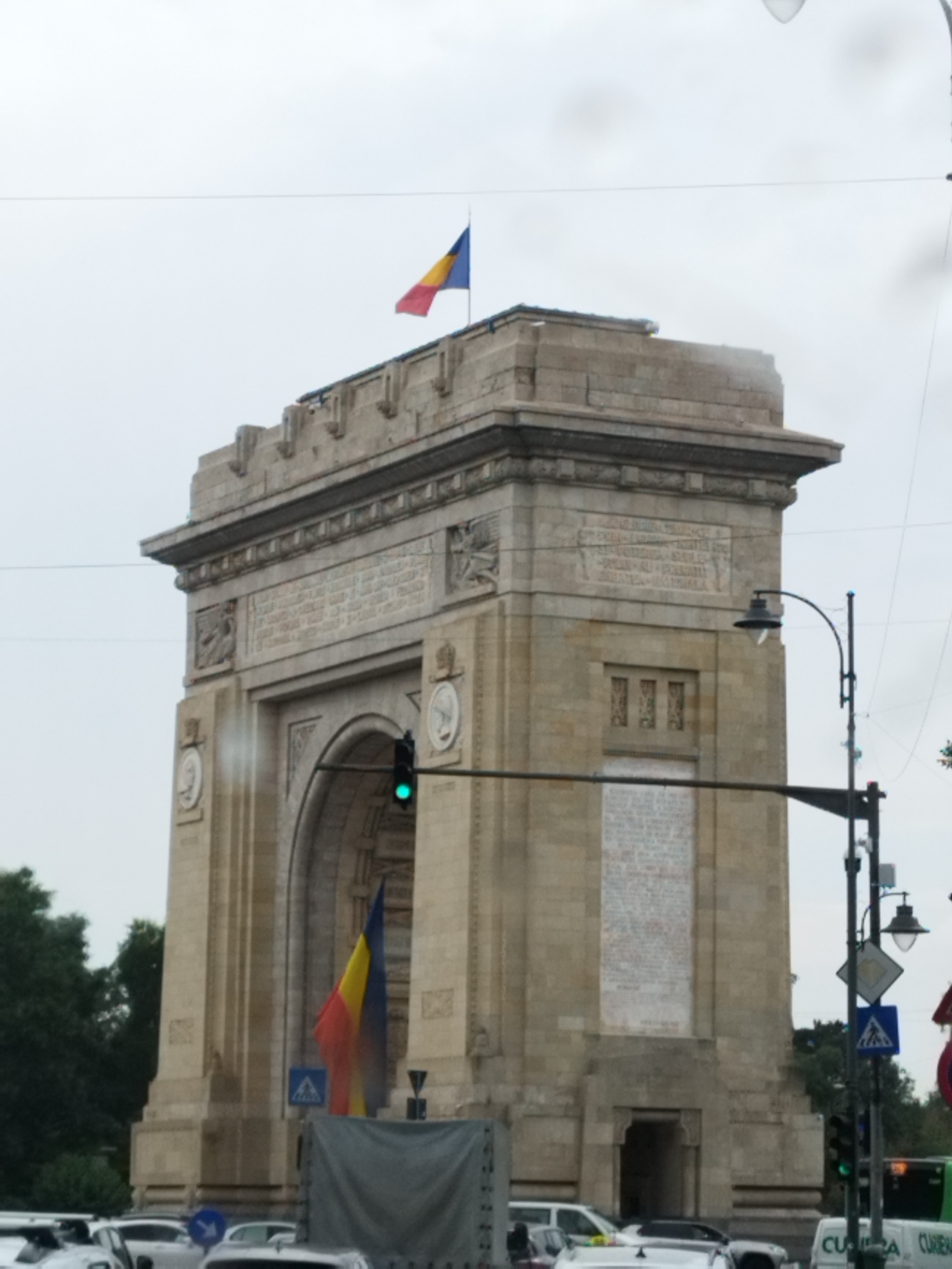 The image shows the Arcul de Triumf in Bucharest, Romania, on a cloudy day. The triumphal arch is made of stone and is decorated with sculptures and inscriptions. A Romanian flag flies from the top of the arch. A traffic light with a green signal is visible on the side of the arch. In the foreground, there are several cars, including a white car, a silver car, and a truck covered with a gray tarp. Trees are visible on the left side of the image, and there are streetlights and traffic signs in the background. The overall scene depicts a street view in Bucharest with the iconic Arcul de Triumf as the central focal point.