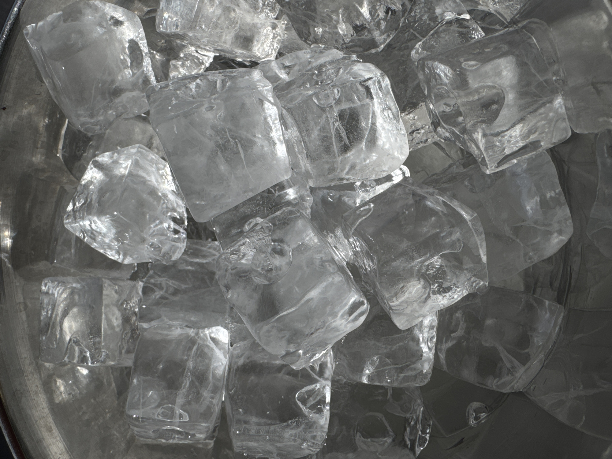 The image is a close-up, overhead shot of a container filled with ice cubes. The container appears to be made of metal. The ice cubes are irregularly shaped and have a transparent to translucent appearance. The lighting is bright, causing highlights and shadows on the surfaces of the ice cubes, enhancing their texture and three-dimensionality. The composition is tightly focused on the ice, with only glimpses of the container visible around the edges. There is no indication of Yerevan, Armenia, or any specific location. No people, subjects, actions, or interactions are depicted.