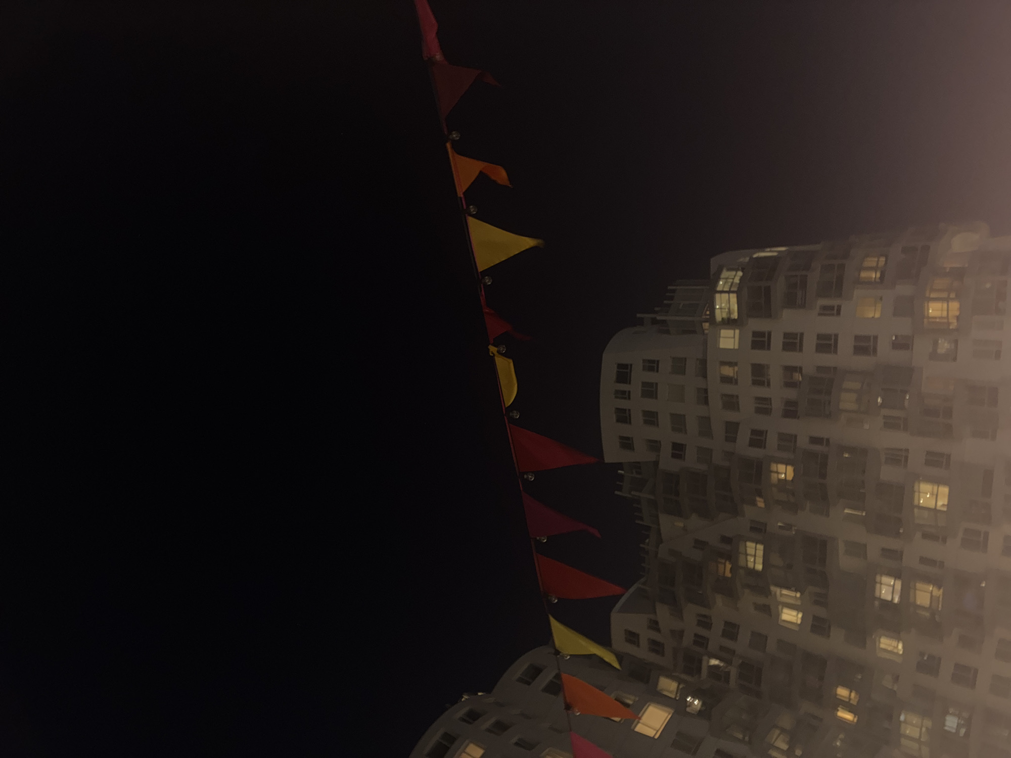 The image shows a night scene with a building and string lights. The building is modern, white, and multi-story with numerous lit windows, suggesting it is either residential or an office space in use after dark. A string of triangular pennant flags is strung diagonally across the foreground, with colors including red, yellow, and orange. The sky is black, indicating it is night. The building style and lighting indicate an urban setting, but additional location detail is impossible without further evidence.