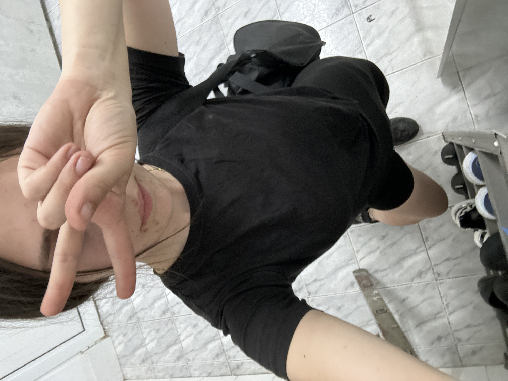 A selfie taken indoors. The subject, a person, is wearing a black outfit and holding up their hand making a "V" sign. A black bag is on the floor behind them. The floor is covered in light gray tile. A weight rack with dumbbells is to the subject's right. The location is not discernible as Resen, Bulgaria.