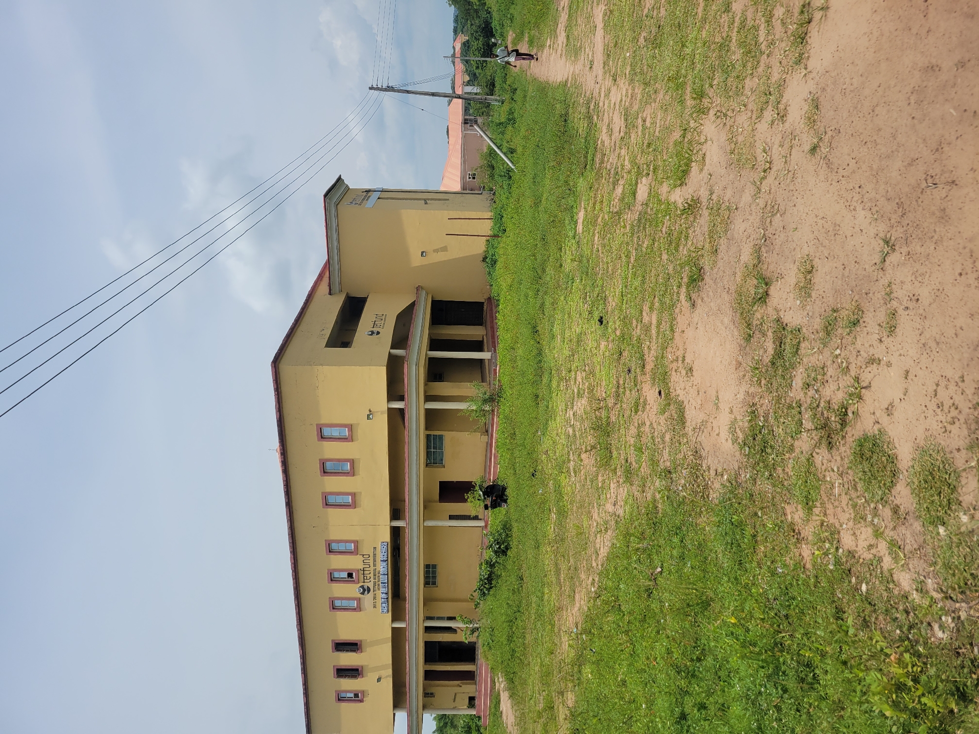 This media file shows a side view of a yellow building with a red roof. The building appears to be an educational institution, as evidenced by the text on its facade. The building has multiple windows and an open-air walkway supported by columns on the first floor. A dirt path runs alongside the building, bordered by green grass and some sparse vegetation. In the distance, more buildings with red roofs are visible, set against a backdrop of green trees and a slightly overcast sky. There are power lines running overhead. A single person, wearing light-colored clothing and a hat, is walking away from the camera on the dirt path in the distance. The lighting suggests it is daytime, with a diffused light indicating either an overcast sky or early morning/late afternoon. The overall scene depicts a somewhat rural or developing area, likely a school or university campus.
Visible text on the building reads: "tetfund" and "2013-2014 TETFUND MEMORIAL UNIVERSITY". The word "TETFUND" appears to be a logo, and the rest is a designation for a period and a specific university.