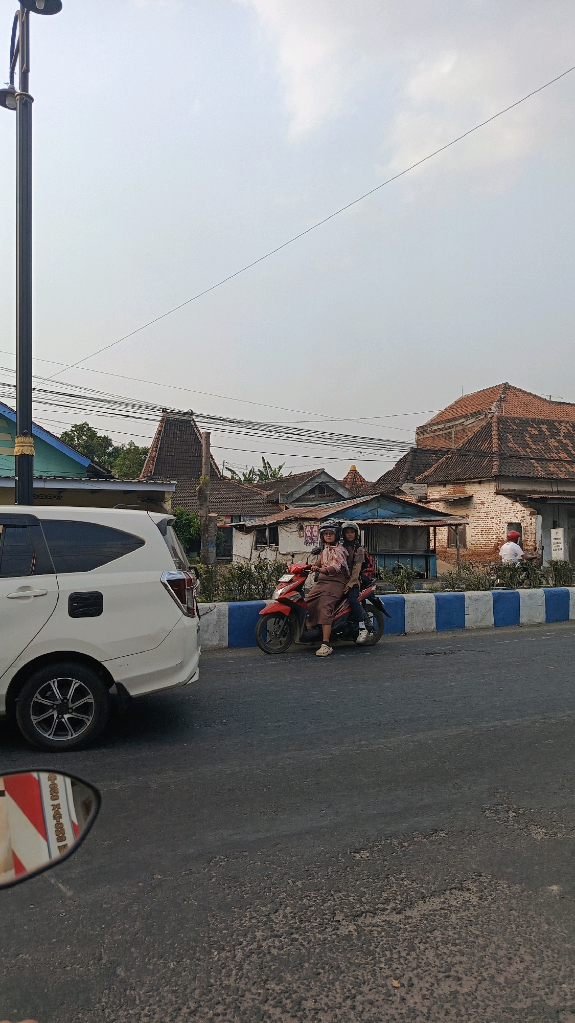 The image depicts a street scene in Mangunsuman, Indonesia. In the foreground, the left side shows the rear of a white SUV. To the right, a red motorcycle is stopped at an intersection. Two individuals, both wearing helmets, are seated on the motorcycle. The person in front appears to be female, wearing a light-colored headscarf and a brown outfit. The person behind them, likely also female, wears a dark headscarf and a beige jacket. They are looking forward, possibly waiting for traffic.
The background reveals a row of buildings with traditional Indonesian architecture, characterized by prominent tiled roofs, some of which are terracotta. The buildings appear to be a mix of residential and commercial structures, with some showing signs of wear. Utility lines are visible strung across the sky. A street lamp stands to the left of the frame. The road surface is asphalt, showing some signs of wear and tear with patches of uneven texture. A blue and white striped barrier runs along the side of the road. The sky is overcast, suggesting either early morning or late afternoon lighting. In the far right background, another motorcycle with a rider wearing a white shirt can be seen. The reflection in the side mirror of the SUV shows a colorful pattern with text, likely from a nearby sign or advertisement.