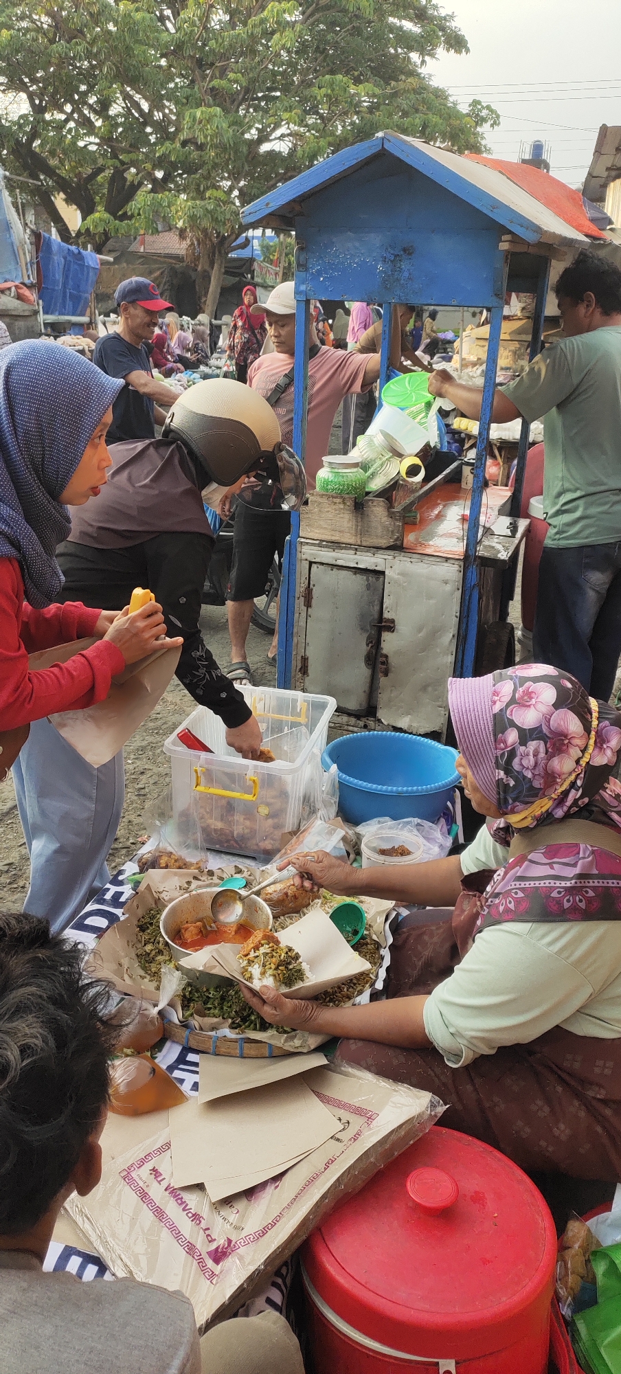 The image depicts a busy outdoor food stall in Karangtowo, Indonesia. In the foreground, a woman wearing a light green shirt and a floral headscarf is serving food from a cart. She is using a ladle to scoop food onto a plate of rice. In front of her, there are several plates of food, some wrapped in paper. To her left, another woman in a red shirt and a blue headscarf is receiving a food packet. A person wearing a helmet is standing behind the cart, and a man in a green shirt is seen on the right side of the frame. The food stall itself is blue and red, with metal cabinets. Behind the stall, there are trees, other stalls, and people in the background, suggesting a lively market atmosphere. The ground is dirt, and the overall scene is illuminated by natural daylight.
