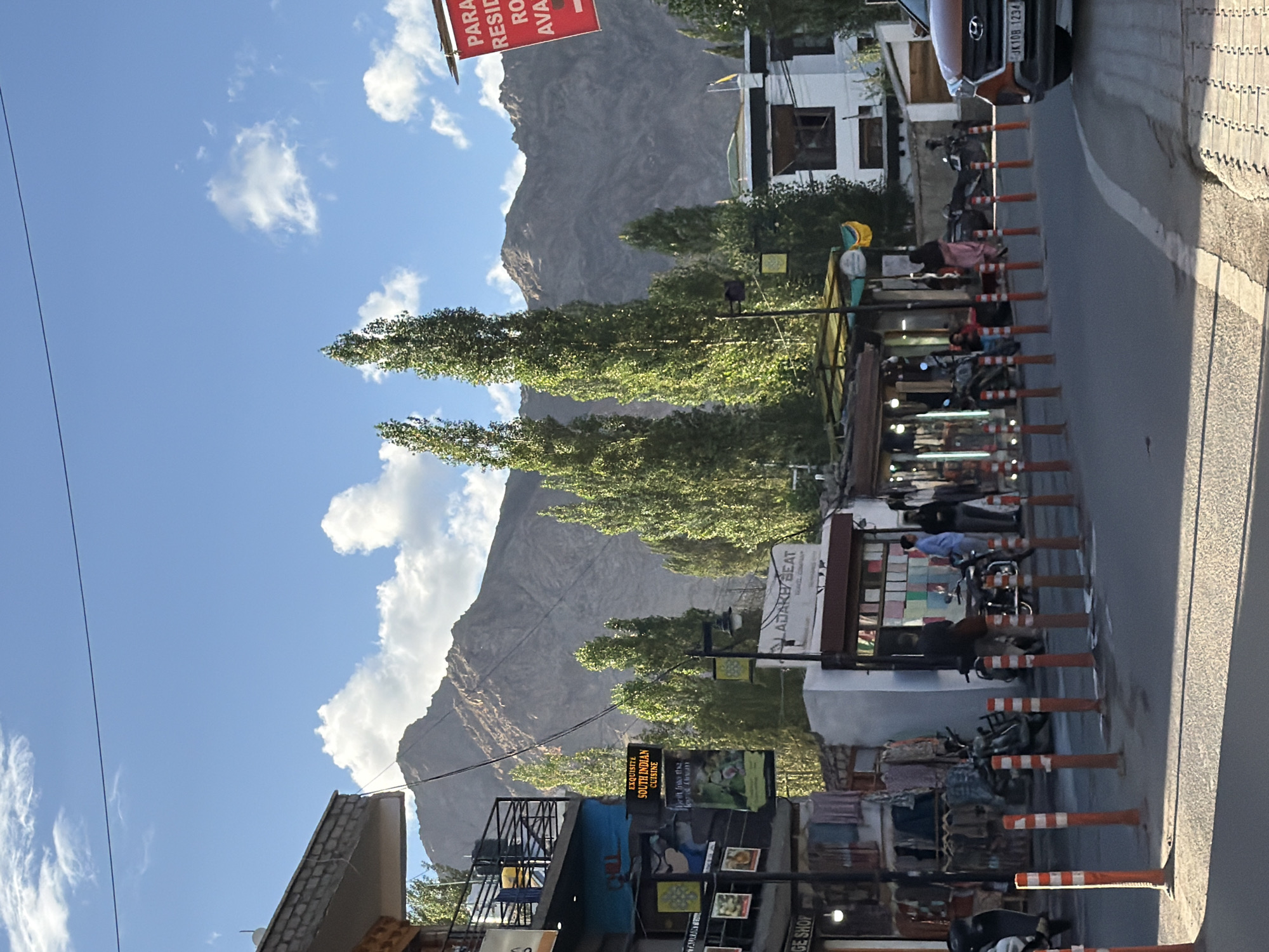A street scene in Leh, India, shows a road lined with shops and vehicles, with a mountainous landscape in the background. The shops display various goods, including clothing and signs for restaurants. Several motorcycles and scooters are parked along the roadside. People are walking and standing in front of the shops. The sky is blue with scattered clouds. The image appears to have been taken during daylight hours.