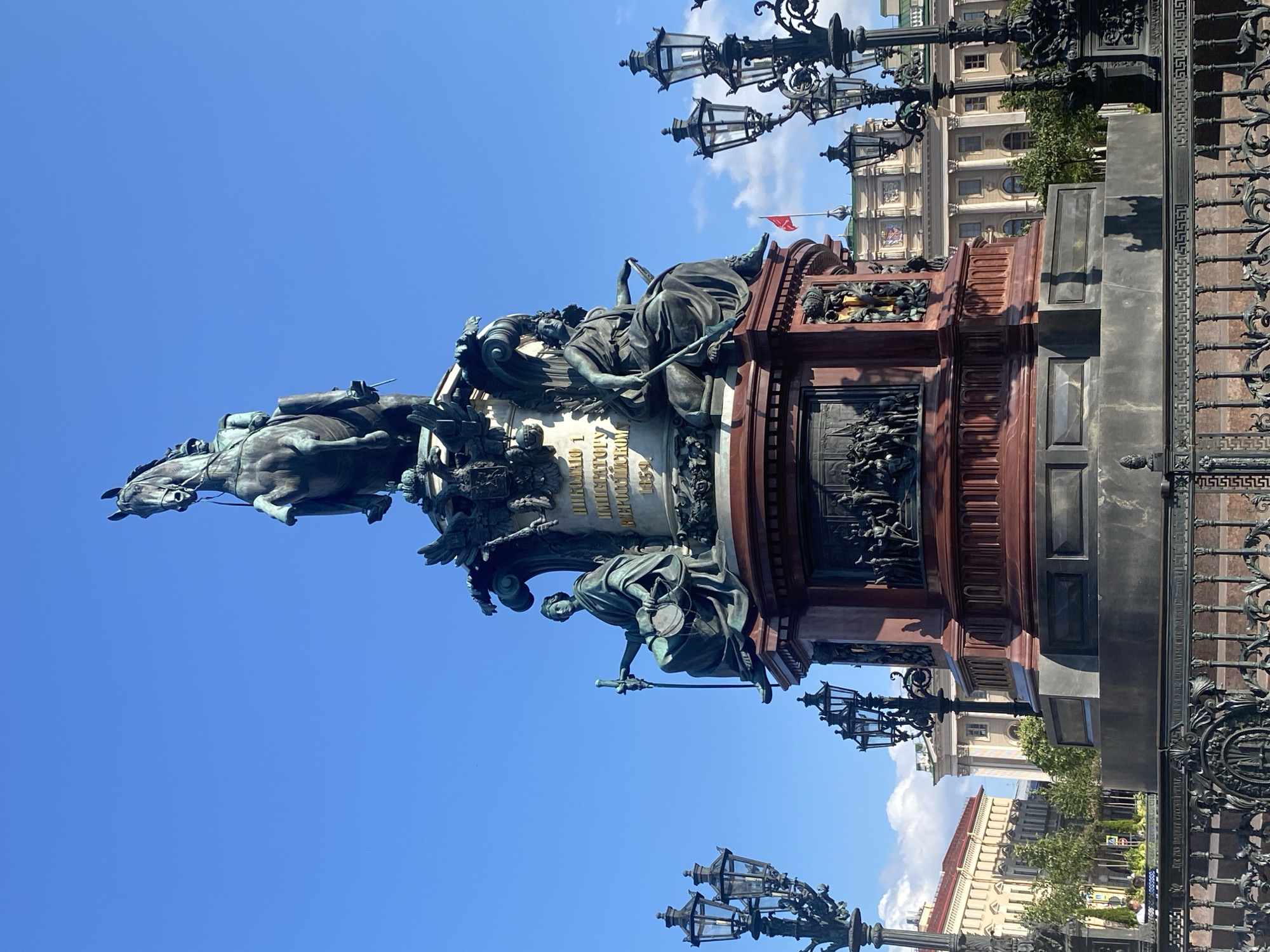 The image shows the Bronze Horseman monument in Palace Square, Saint Petersburg, Russia. The monument features a large equestrian statue of Peter the Great atop a granite pedestal. Surrounding the monument are ornate lampposts and in the background, stately classical buildings of Saint Petersburg are visible under a clear blue sky with scattered clouds. The monument itself is dark bronze, while the pedestal is a deep red. The details of the figures on the pedestal, the horse, and Peter the Great are clearly discernible, as are the intricate metalwork of the surrounding fence and lampposts. The scene is illuminated by daylight, casting shadows on the monument and the surrounding area. No people are prominently featured in the image.