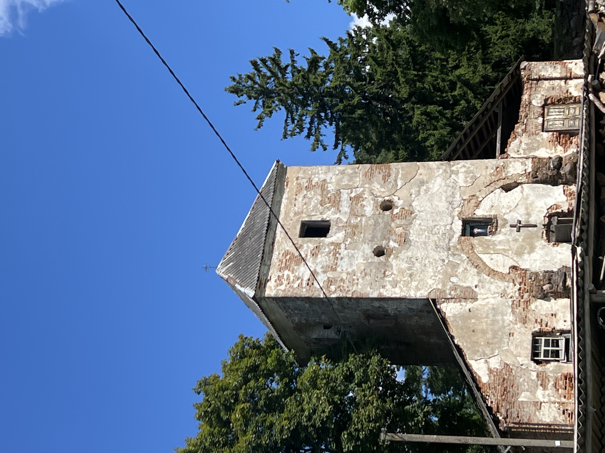 The image captures the exterior of a partially ruined stone and brick structure, possibly a former church or chapel, under a clear blue sky in Norviliškės, Lithuania. The building exhibits significant wear and tear, with exposed brickwork and crumbling plaster. A corrugated metal roof covers a portion of the structure, and an arched entrance with visible brick detailing is present. Small windows, some with panes and others open, dot the facade. A cross is visible above one of the windows, and another smaller cross is positioned near an arched recess. Mature trees, including a tall evergreen, surround the building, suggesting a rural or semi-rural setting. A dark electrical wire diagonally bisects the upper portion of the image. The overall impression is one of historical decay and abandonment.