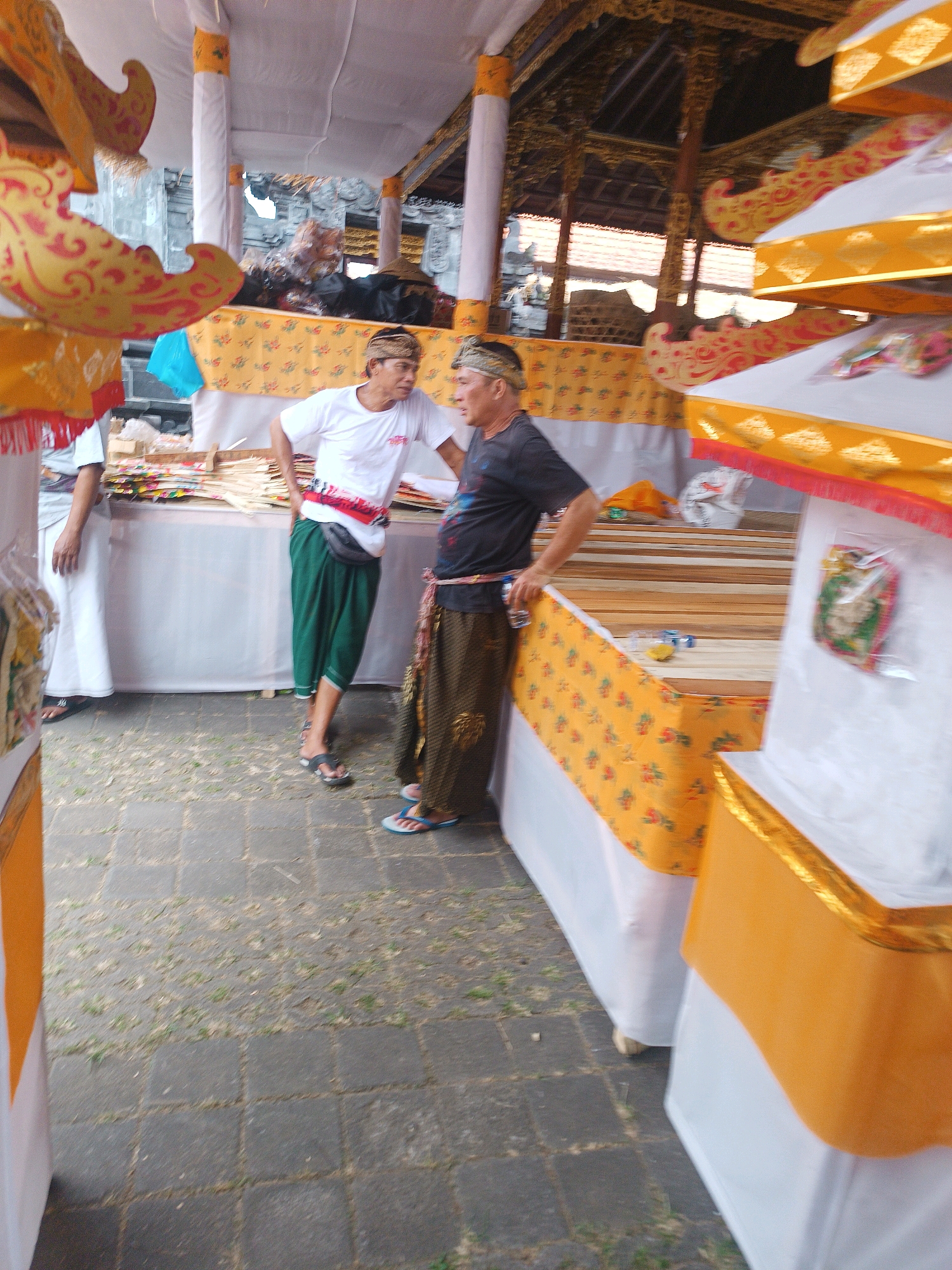 Two adult males, appearing to be of Indonesian descent, are standing outdoors in what seems to be a market or a festive gathering. One male is wearing a white t-shirt, green pants, and a black fanny pack, with his left hand on his hip. The other male is wearing a dark t-shirt, brown patterned pants, and a red sash, holding a bottle of water. They are positioned near stalls adorned with yellow and red fabric, possibly for a ceremony or sale. In the background, traditional Indonesian architecture with ornate golden decorations is visible. The ground is paved with dark square stones. The overall atmosphere suggests a cultural event in Selemadeg, Indonesia.