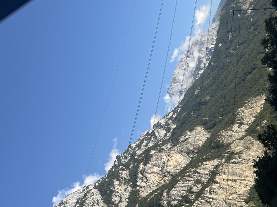 The image displays a mountainous landscape near Schattdorf, Switzerland, characterized by steep, rocky formations and dense coniferous forestation under a clear blue sky with scattered white clouds. A prominent mountain face with visible rock striations and green tree coverage occupies the left and center of the frame. In the distance to the right, a higher, more rugged peak with light-colored rock, possibly snow-dusted, is partially shrouded by clouds. Multiple dark, parallel power lines traverse the mid-section of the image horizontally. Dark green foliage, likely tree tops, is visible along the bottom edge of the frame. No individuals, specific actions, or additional discernible man-made structures are present.