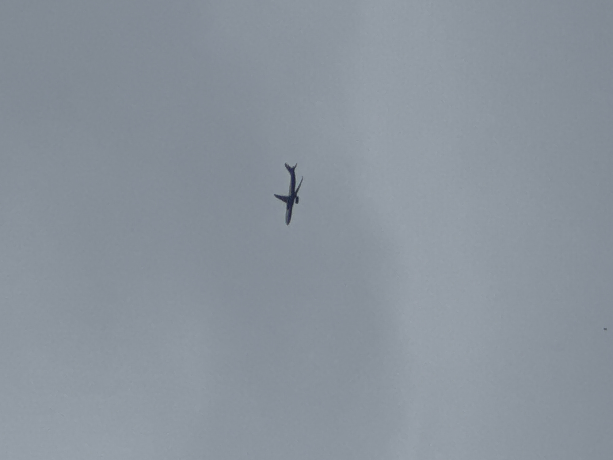 A passenger airplane is flying in the sky above Pa Khlok, Thailand. The sky is overcast. A small dark object is also visible in the lower right quadrant of the frame.
