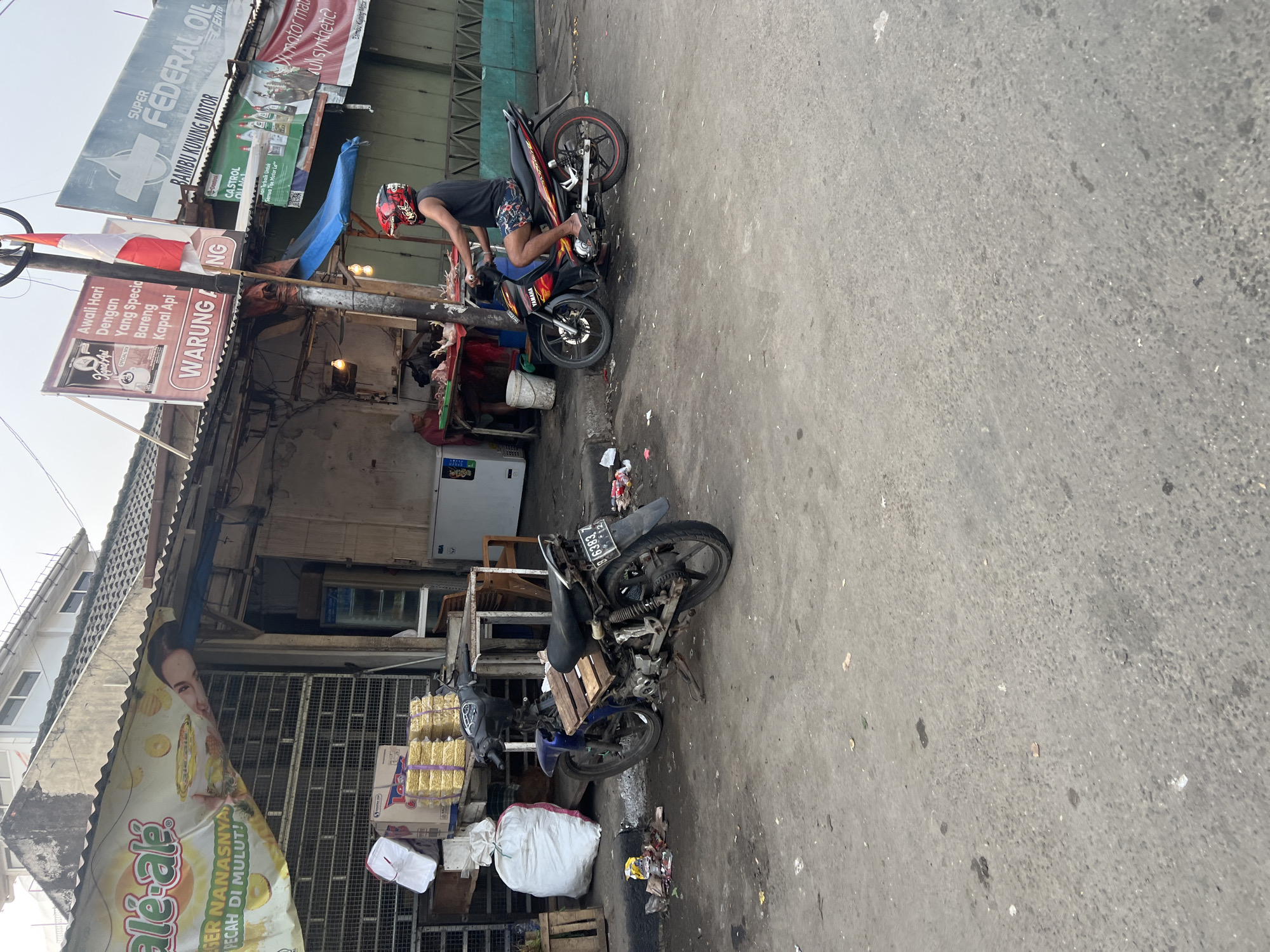 In Citeureup, Indonesia, a street scene unfolds with two motorcycles parked on a dusty asphalt road in front of a row of small shops. The shop on the left displays a banner with the text "ale-ale" and images of lemons. Next to it, stacks of goods wrapped in yellow plastic are visible. A person in a red helmet and a dark sleeveless shirt is seated on one of the motorcycles, which is positioned to the right. Another motorcycle, a darker color, is parked on the left. The environment appears somewhat cluttered, with trash scattered on the ground. In the background, a person is seen near a table laden with poultry, suggesting a market or food stall. Billboards with various advertisements are attached to the buildings. The lighting indicates it is daytime.