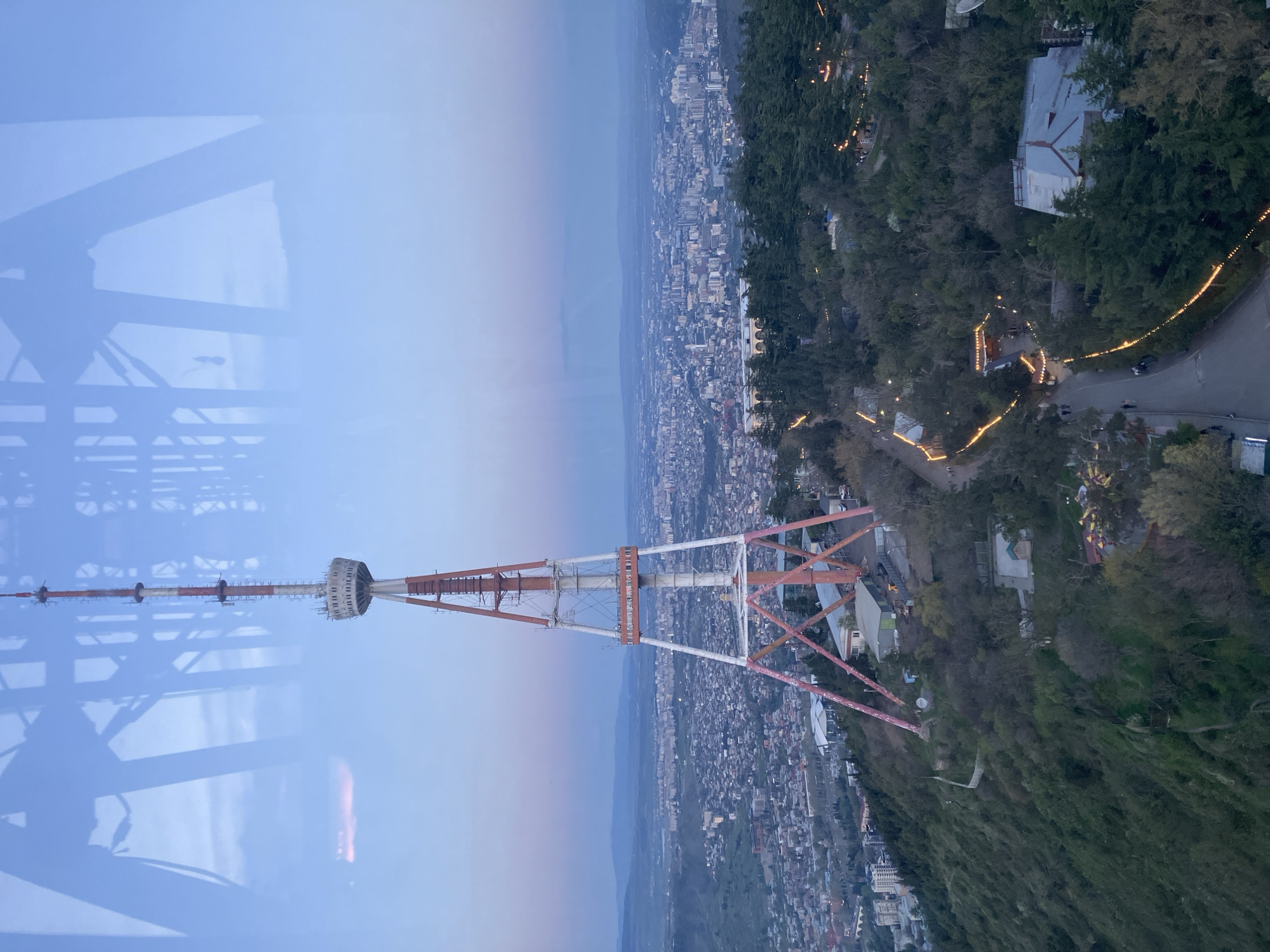 The image captures a panoramic view of Tbilisi, Georgia, during dusk or early evening. The scene is dominated by the prominent Tbilisi TV Tower, a red and white structure featuring a circular section towards its upper portion, visible in the mid-ground.
The foreground and mid-right of the image consist of a densely forested hillside, through which winding roads are illuminated by numerous string lights, suggesting the presence of outdoor venues or establishments. To the left and stretching into the distance is the expansive cityscape of Tbilisi, with a multitude of buildings and streetlights beginning to illuminate the urban landscape. The sky above is a soft blue, gradually fading into a lighter hue near the horizon, indicative of twilight.
The upper left section of the photograph shows reflections on a glass surface, suggesting the image was captured from inside a building or an enclosed viewing platform. These reflections include indistinct structural elements. The overall visibility is clear, and the atmosphere appears calm. There are no identifiable people or visible text in the image.