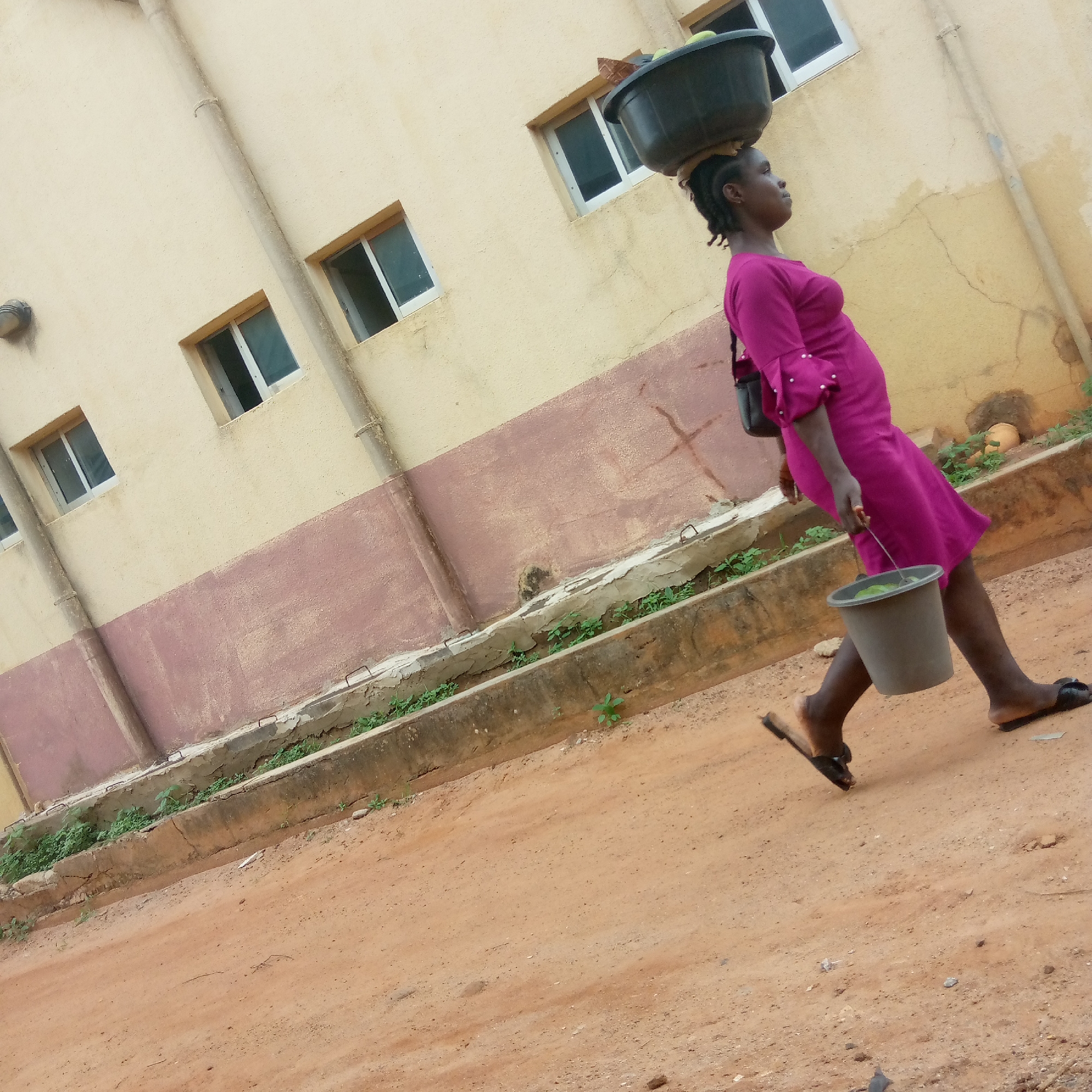 A woman in a magenta dress walks on a dirt path beside a building. She carries a large black basin on her head and a gray bucket in her right hand. The building is two-toned, with a beige upper section and a maroon lower section. It has several windows with white frames. The area appears to be Ukwu-Abwa, Nigeria, based on the photographic style and potential clues like architecture and typical garb. The woman's task appears to be water transportation based on the buckets/basins.